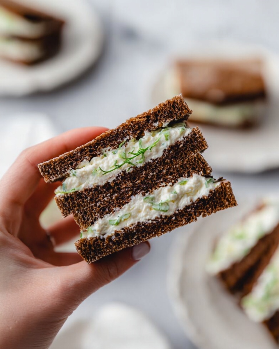 A close-up of a woman's hand holding a sandwich made of two dark brown slices of bread with a creamy white filling that has small green bits inside. The sandwich is cut into two small triangular pieces stacked on each other, showing two layers of bread and one layer of filling in between. The background has a white marbled texture and blurred white plates with more sandwiches on them. Photo taken with an iphone --ar 4:5 --v 7