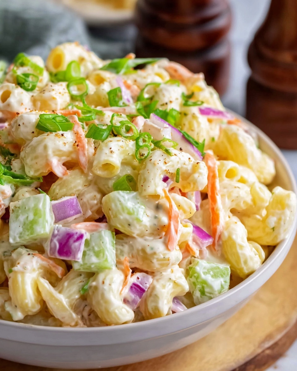 This image shows a close-up view of a creamy pasta salad in a white bowl on a white marbled surface. The salad consists of three layers: the base layer is pale yellow macaroni pasta with a smooth texture, topped with small pieces of crunchy celery in light green, thin orange carrot strips, and finely chopped red onion which adds spots of purple. Bright green chopped scallions are scattered on top, adding a fresh touch. The whole dish is coated in a thick, white creamy dressing making the ingredients look shiny and mixed together. In the background, blurred dark pepper grinders and other kitchen items are faintly visible. Photo taken with an iphone --ar 4:5 --v 7