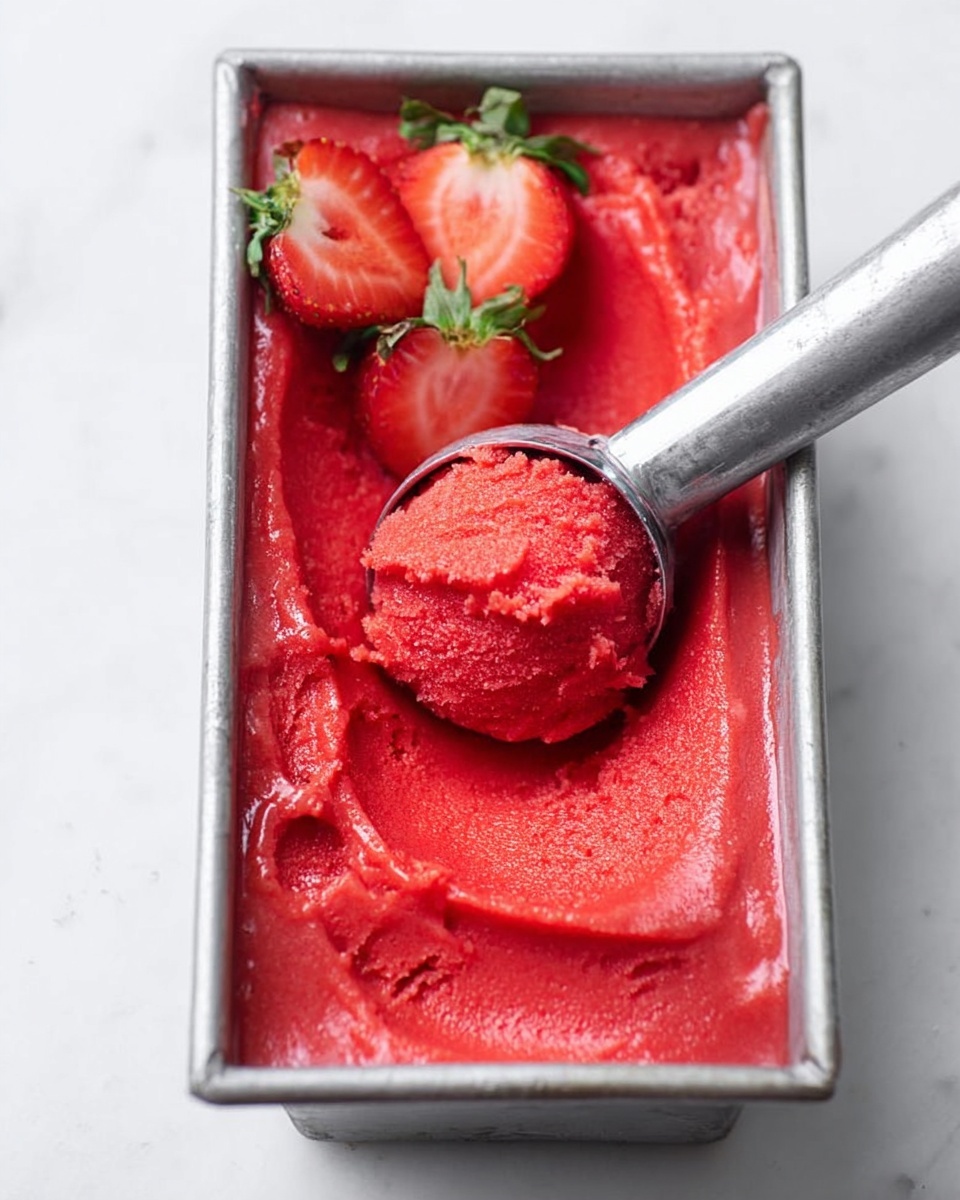 The image shows a metal rectangular container filled with smooth, bright red strawberry sorbet that has a soft and slightly textured surface. The sorbet fills the container fully and has visible swirls and uneven spatula marks on top. Two halved fresh strawberries, showing their juicy red inside with green leafy tops, are placed on the bottom right corner of the sorbet inside the container. The container is placed on a white marbled surface. photo taken with an iphone --ar 4:5 --v 7