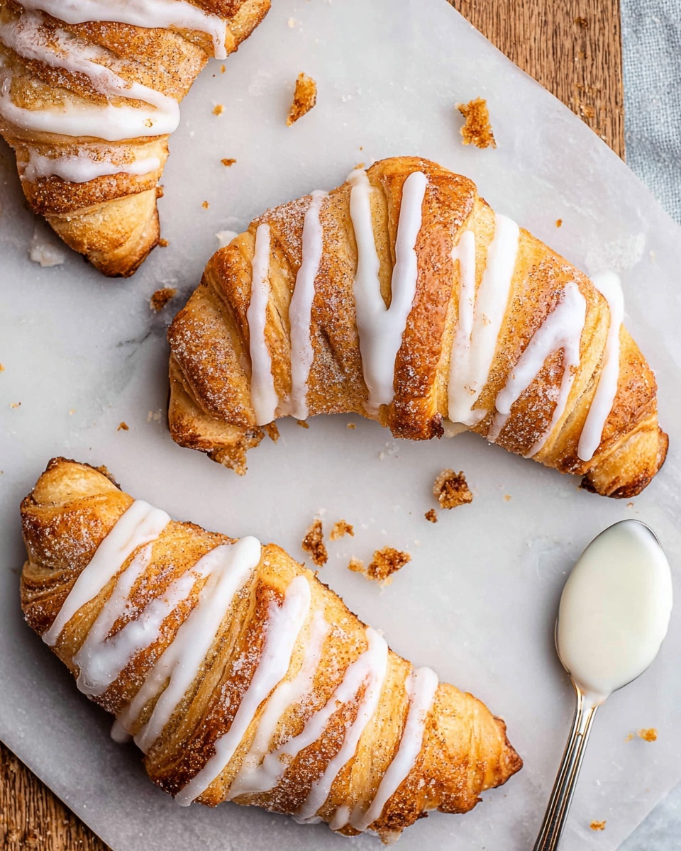 Three golden-brown crescent-shaped pastries lie flat on a white marbled surface, each with a visible layer of cinnamon sugar filling inside and topped with thin white icing stripes drizzled across the top. The pastries have a slightly crisp texture with some browned edges, and small crumbs are scattered around them. In the top right corner, a small spoon holds a bit of white icing spilling onto the white marbled surface. photo taken with an iphone --ar 4:5 --v 7