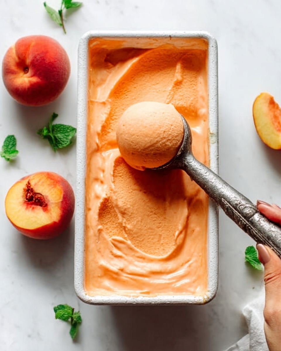The image shows a rectangular white metal container filled with smooth, creamy orange peach ice cream. A woman's hand is holding a vintage-style metal scoop lifting a round ball of the ice cream from the container, with soft swirls and texture visible inside. Around the container, there are a few whole fresh peaches and small green mint leaves scattered on a clean white marbled surface. The lighting is soft and natural, highlighting the ice cream’s velvety texture and bright color. Photo taken with an iphone --ar 4:5 --v 7