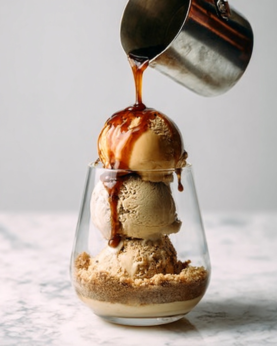 The image shows a clear glass dessert cup with three scoops of ice cream stacked on top of each other. The bottom layer inside the glass is a light brown crumbly layer, almost like crushed cookies or nuts. Above this are three smooth and creamy ice cream scoops in shades of beige to light brown. A metal container held by a woman’s hand is pouring a dark brown sauce over the top scoop, which drips down the sides of the ice cream. The background and surface have a white marbled texture. Photo taken with an iphone --ar 4:5 --v 7