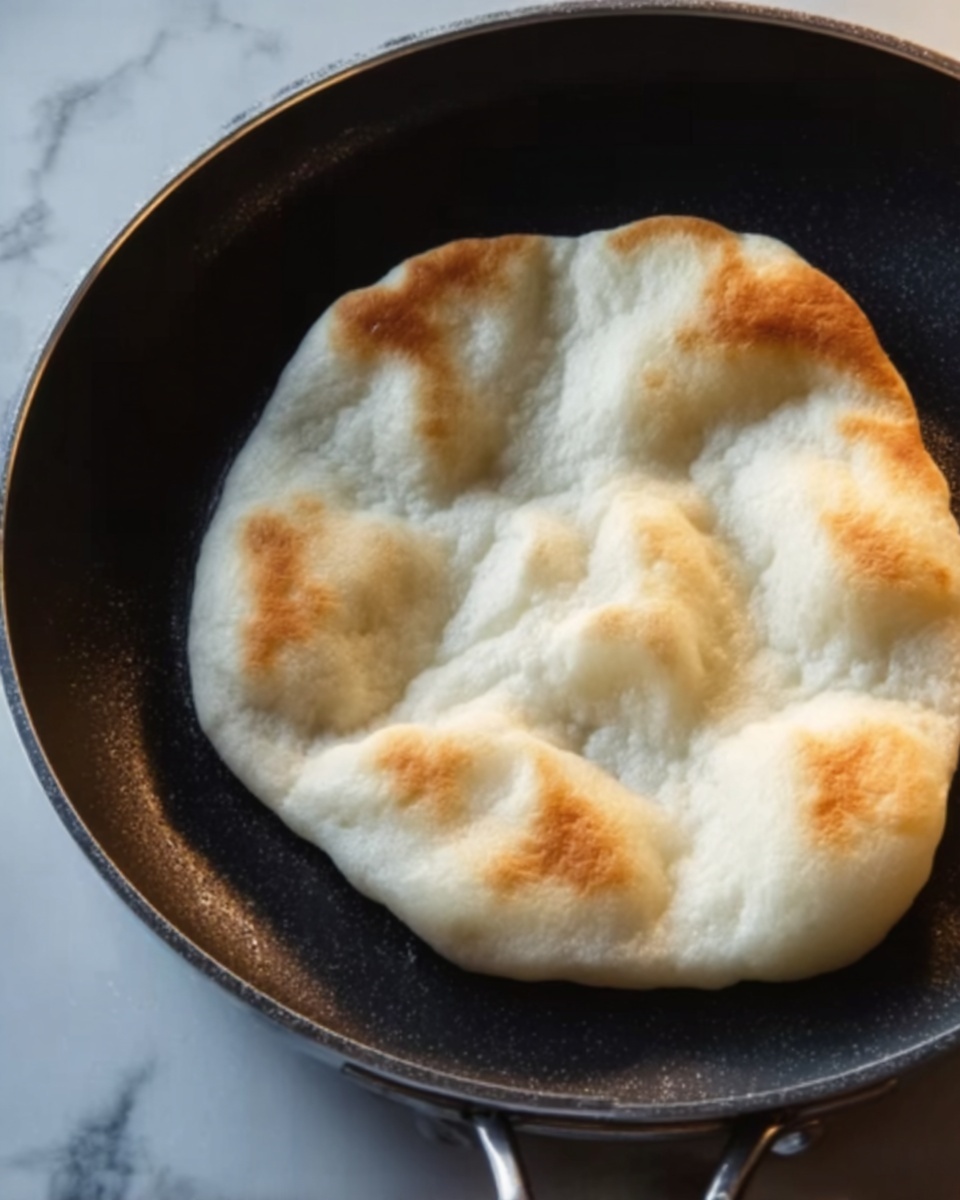 A close-up of a dark frying pan with a white puffed flatbread cooking inside. The flatbread has four raised bubbly sections with a soft, fluffy texture and a slightly golden, crisp edge. The pan sits on a white marbled surface that contrasts with the dark pan. The lighting highlights the smooth surface of the bread and the slight air pockets. Photo taken with an iphone --ar 4:5 --v 7