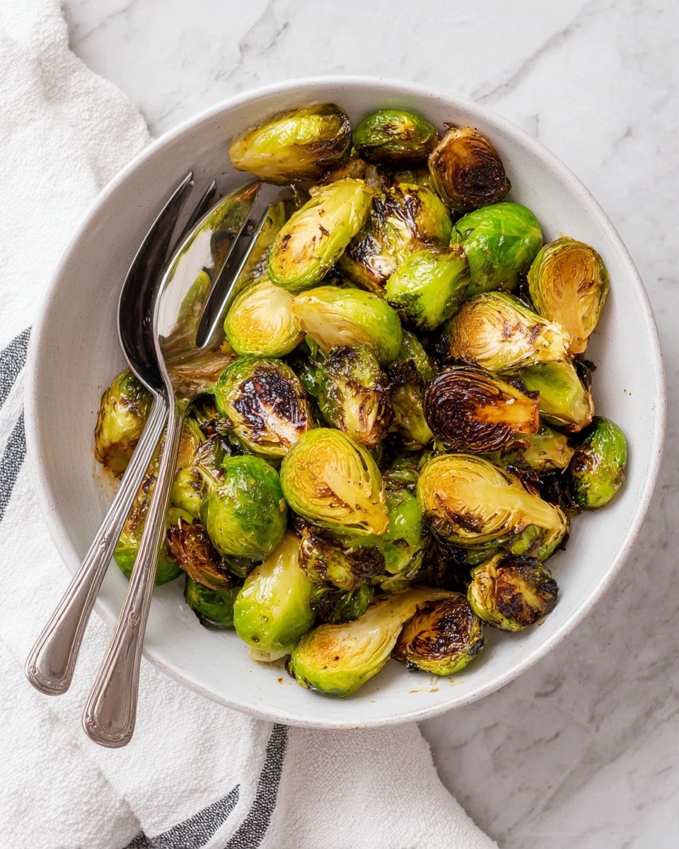A white bowl filled with roasted Brussels sprouts, each cut in half showing light green inner layers with browned, crispy edges and charred spots. The Brussels sprouts are spread inside the bowl, with colors ranging from bright green to golden yellow and dark brown from roasting. Two silver spoons, one solid and one with slots, rest inside the bowl on the left side, partially covered by the sprouts. The bowl sits on a white marbled surface with a white cloth with dark stripes partially visible at the bottom left. Photo taken with an iphone --ar 4:5 --v 7