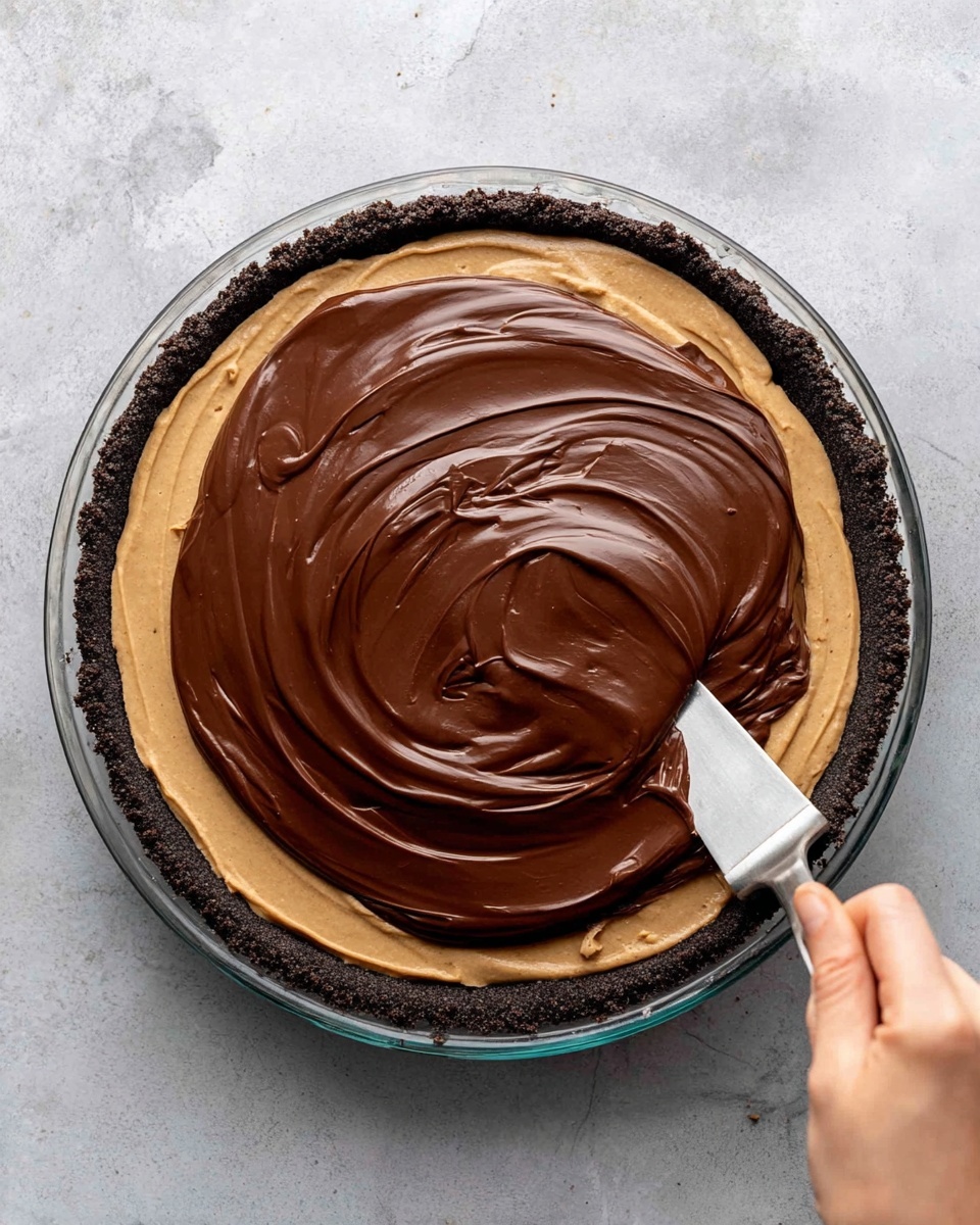 A close-up top view of a pie in a glass pie dish placed on a white marbled surface. The pie has three layers: the bottom is a thick, dark crumbly crust that lines the dish edges, the middle layer is a smooth, light brown filling spread evenly, and the top layer is a glossy, rich dark chocolate ganache being spread with a flat metal spatula held by a woman's hand in the lower right corner. The ganache has soft swirls showing the spreading motion. Photo taken with an iphone --ar 4:5 --v 7