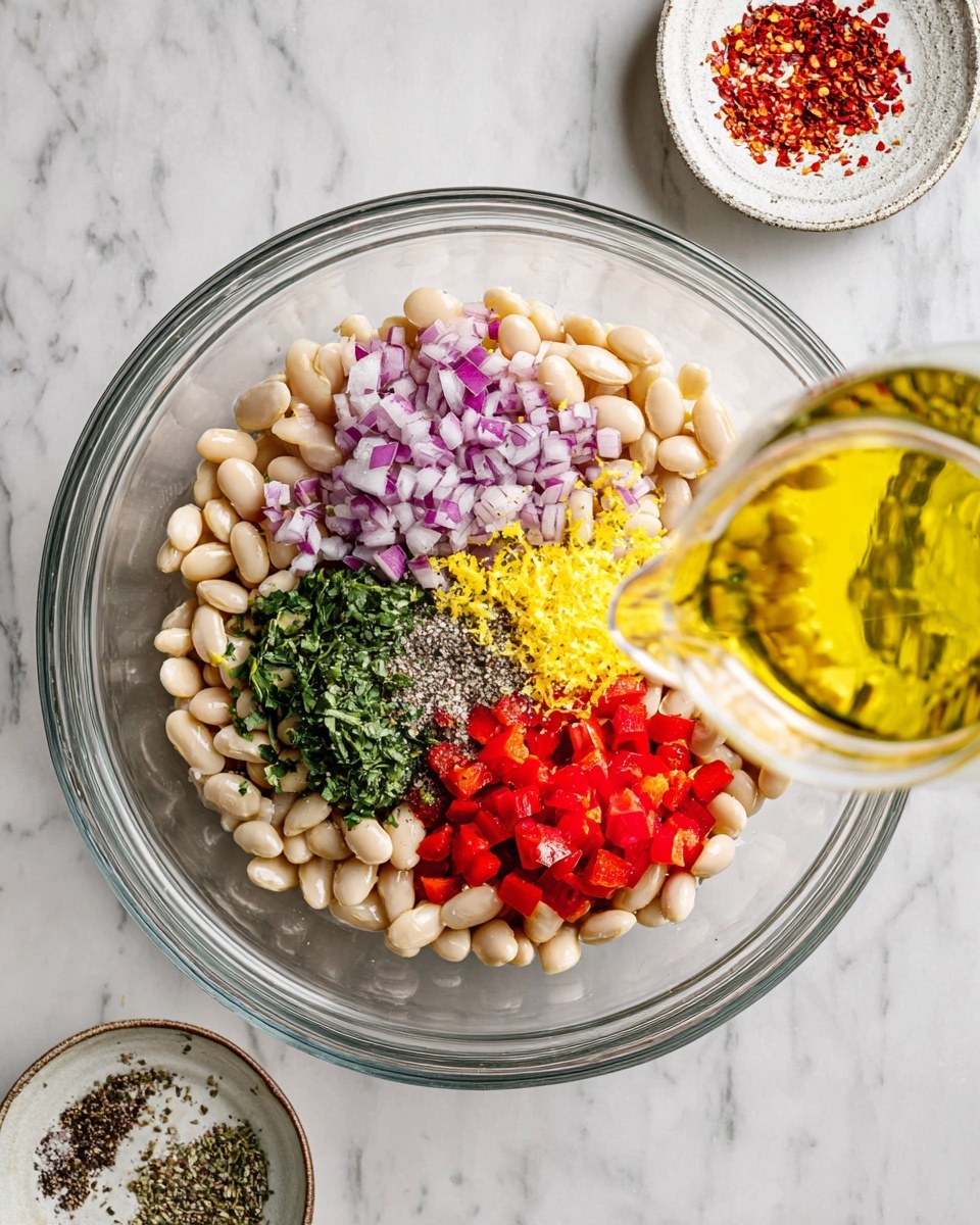 A clear glass bowl sits on a white marbled surface filled with one layer of white beans spread around the edges. Inside the beans are grouped ingredients arranged in small piles: finely chopped purple onions on the left, chopped bright red peppers on the bottom right, bright yellow lemon zest in the middle, chopped green herbs above the zest, and a small mix of white salt and black pepper near the top center. From the right side, a transparent glass container pours a golden yellow liquid over the beans. Nearby, on the white marbled surface, there are two small round dishes—one with red chili flakes and the other with salt and black pepper. Photo taken with an iphone --ar 4:5 --v 7