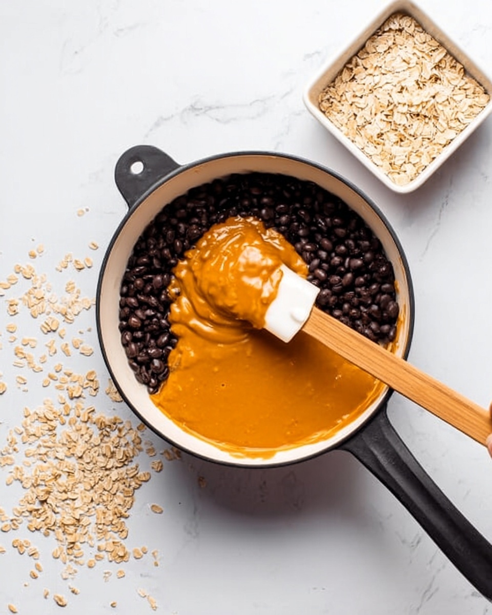 A white saucepan with black handles is placed on a white marbled surface. Inside the saucepan, there are two main layers: on one side, small black beans fill about half the pot, showing a smooth, slightly shiny texture, and on the other side, an orange thick sauce or melted cheese is being spread over the beans; a woman's hand holds a white spatula with a wooden handle just above the pot, touching the orange layer. To the top right of the saucepan, a white square bowl contains light beige rolled oats, some scattered on the marbled surface near the pot. The image is bright, clean, and focused on the pan and its contents. photo taken with an iphone --ar 4:5 --v 7