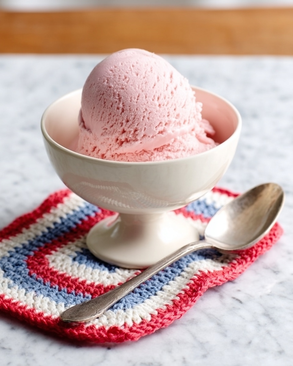A scoop of pink ice cream sits in a white pedestal bowl with smooth texture and a slight sheen, showing creamy softness. The bowl is placed on a small, colorful crocheted mat featuring red, white, and blue stripes with a zigzag edge. Beside the bowl, on the white marbled surface, lies a shiny silver spoon with a gently curved handle resting flat. The photo taken with an iphone --ar 4:5 --v 7