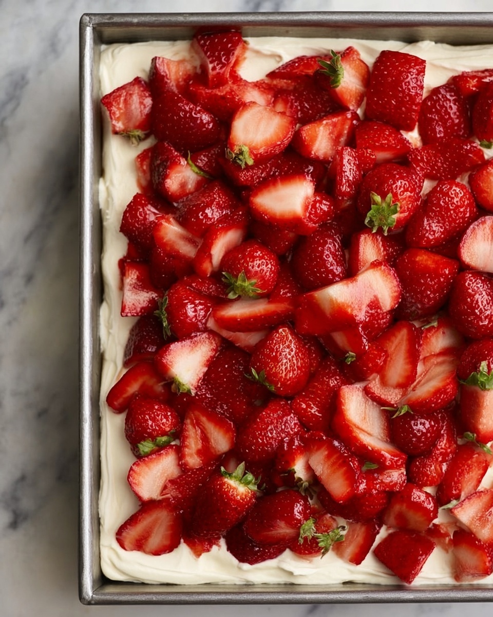 The image shows a rectangular metal pan filled with a dessert that has two layers. The bottom layer is a thick and creamy white base with a smooth and slightly fluffy texture, spread evenly across the pan. The top layer is covered with many fresh red strawberries, some whole with green tops and others sliced to show their juicy inside, arranged closely together. The dessert is placed on a white marbled surface. Photo taken with an iphone --ar 4:5 --v 7