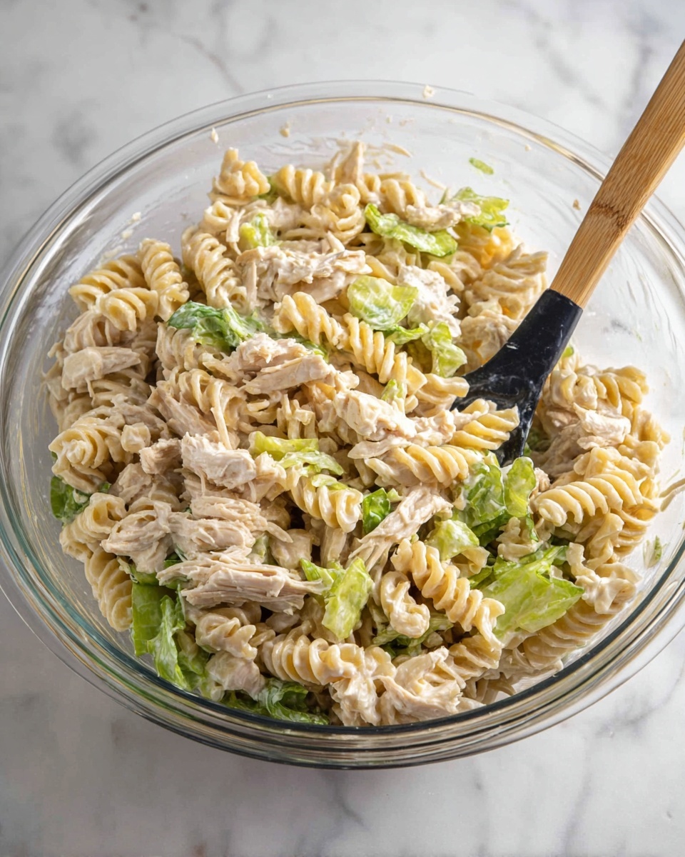A clear glass bowl holds a mixed pasta salad with three main layers: creamy light beige sauce coating twisted rotini pasta, shredded shredded light tan cooked chicken mixed evenly throughout, and small pieces of bright green leafy lettuce scattered on top and inside. A black spoon with a wooden handle is placed inside the bowl on the right side, resting over the pasta mixture. The bowl is set on a white marbled surface. Photo taken with an iphone --ar 4:5 --v 7