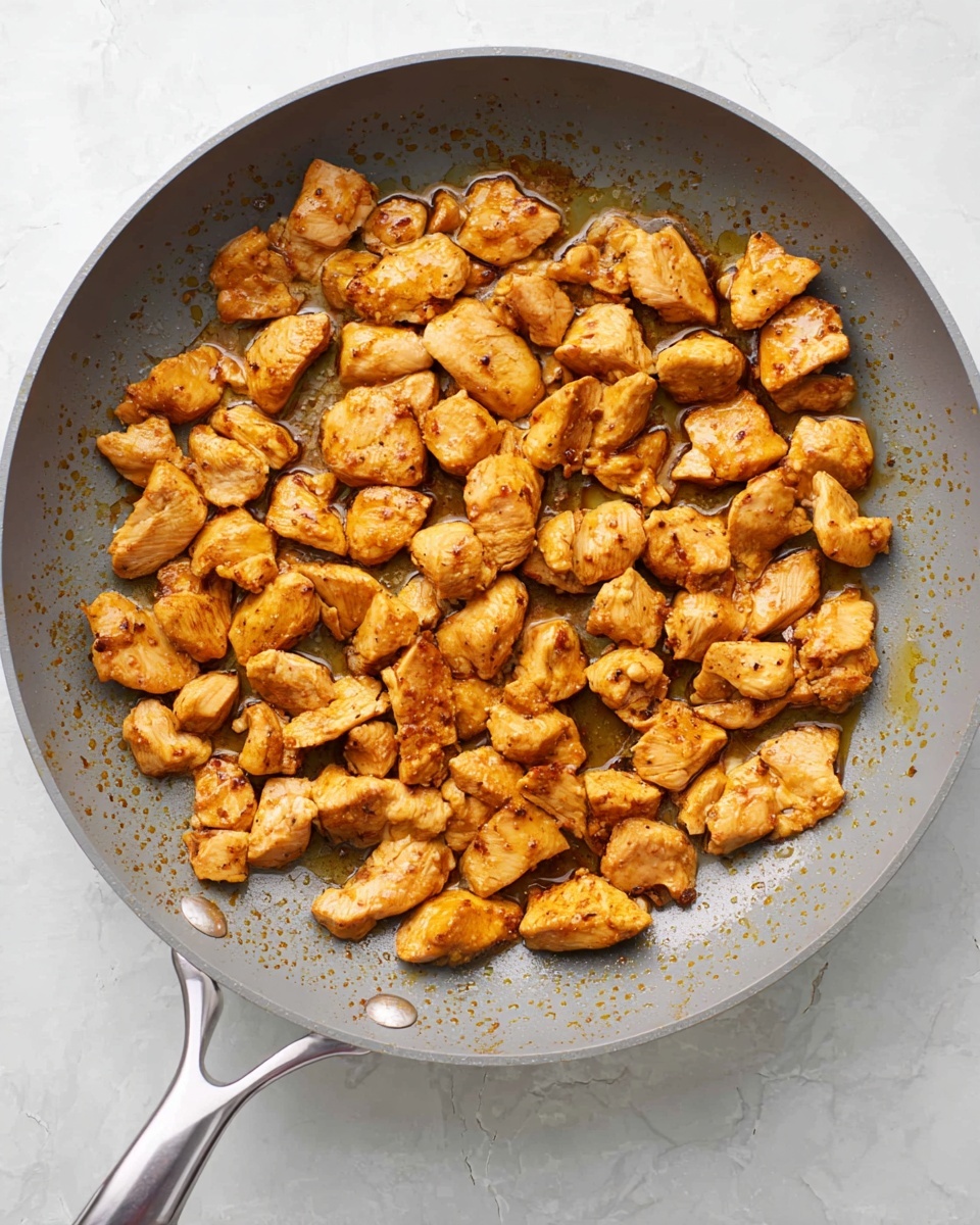 A round gray frying pan filled with many small pieces of cooked golden brown chicken, evenly spread out with some bits showing a slightly darker sear. The pan sits on a white marbled textured surface, and the chicken pieces have a moist and tender look with some oil glistening around them. The pan handle is silver and shiny, positioned to the left side of the image. photo taken with an iphone --ar 4:5 --v 7