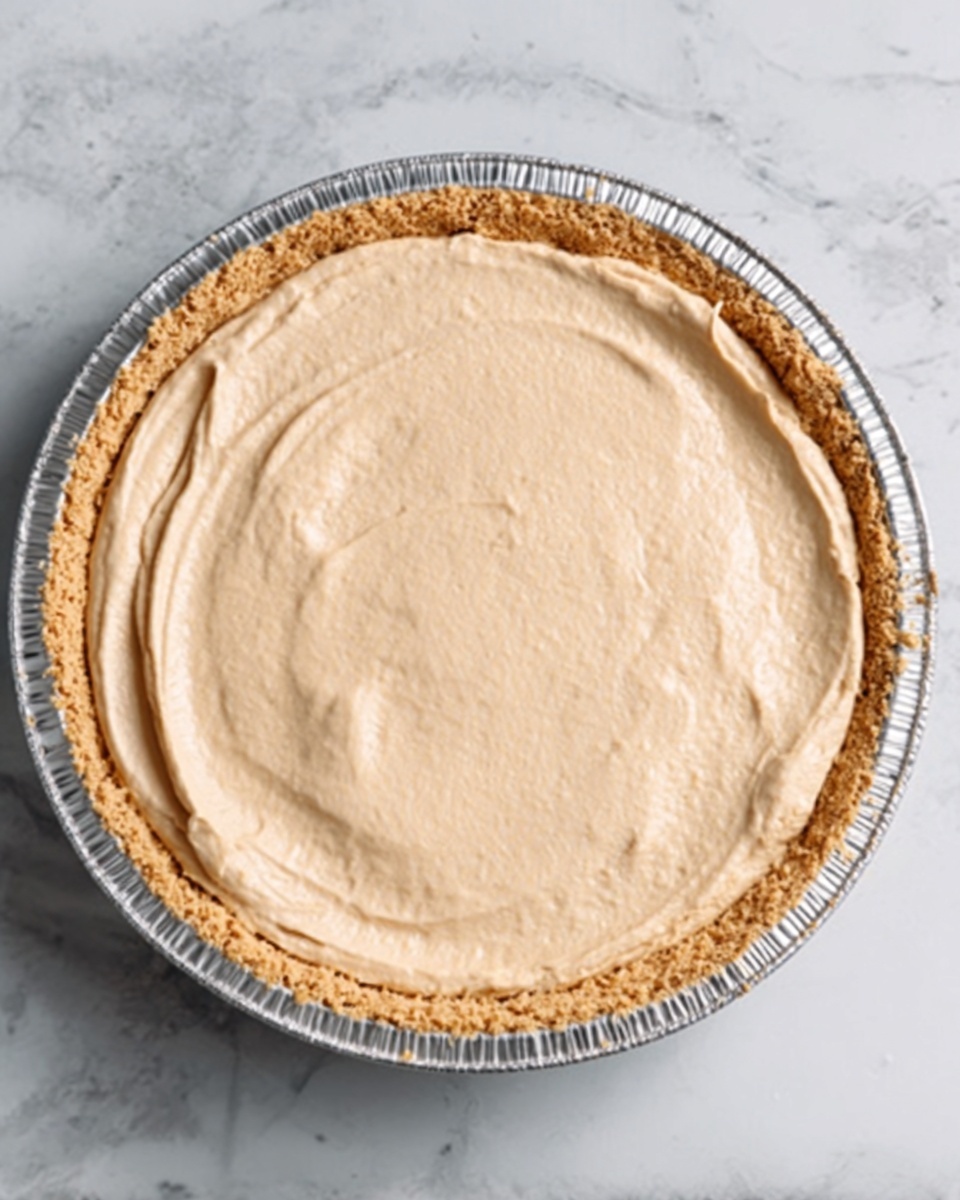The image shows a close-up of a round dessert in a foil pie tin. The dessert has one smooth, creamy top layer with a light beige color. The base looks crumbly and pale brown, with a slightly thicker edge around the pie. The pie tin sits on a white marbled surface. The light makes the top look soft and slightly shiny. photo taken with an iphone --ar 4:5 --v 7