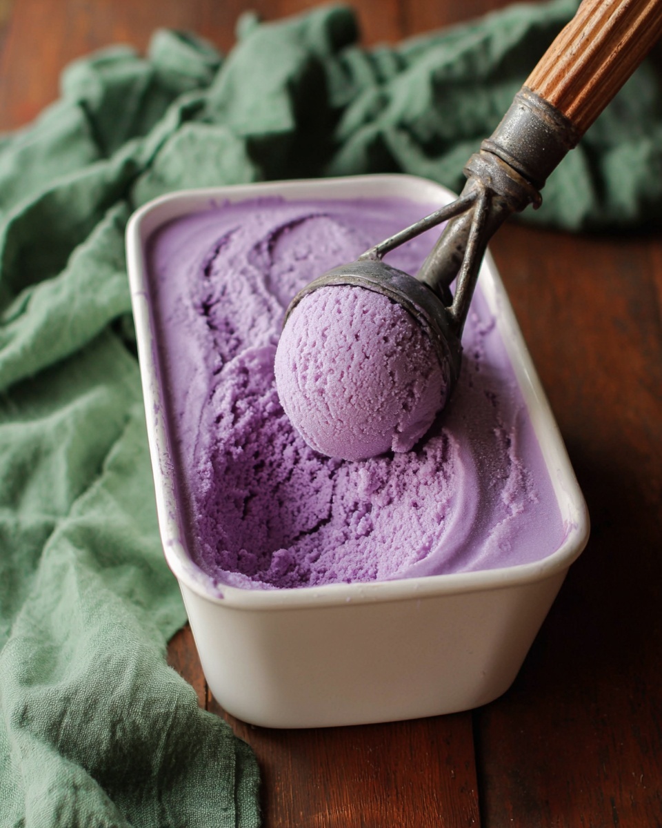 A white tall container filled with one layer of creamy purple ice cream, with a scoop taken out, showing a soft texture. An old metal ice cream scooper with a wooden handle is lifting a rounded scoop of the purple ice cream from the container. In the background, a blurred green cloth lies on a wooden surface. photo taken with an iphone --ar 4:5 --v 7