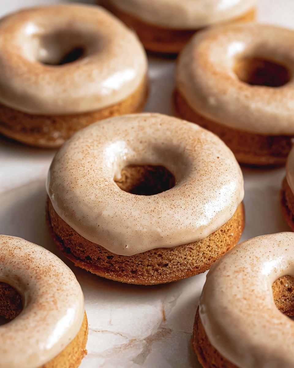The image shows several round donuts placed on a white marbled surface. Each donut has two layers: a thick, dense, golden-brown base and a smooth, light beige glaze covering the top layer. The glaze appears slightly shiny with a dusting of fine light brown powder, likely cinnamon or sugar, giving a textured look. The donuts are closely arranged, with the focus on the center donut which looks slightly raised above the others. The overall scene is warm and inviting with soft lighting highlighting the textures. photo taken with an iphone --ar 4:5 --v 7