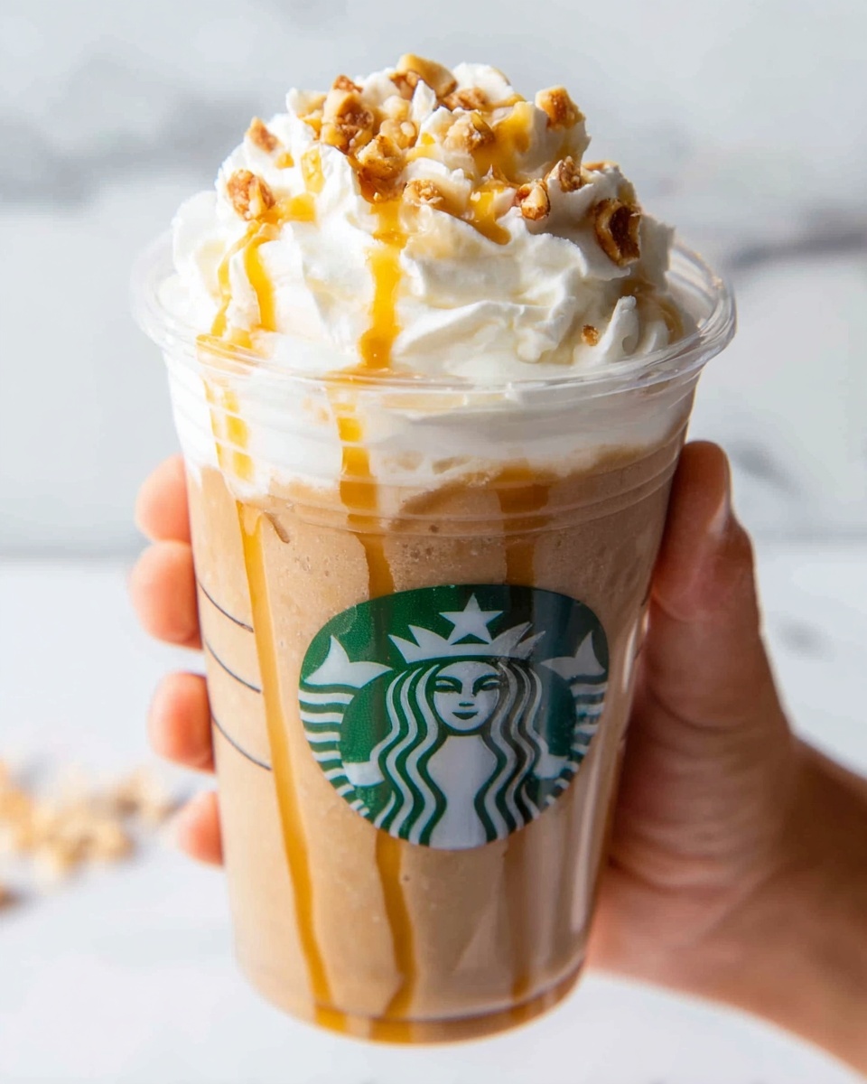 This image shows a clear plastic cup filled with a light brown blended coffee drink. The drink has thick caramel syrup drizzled inside the cup, flowing down the sides in uneven lines. The top layer is a generous swirl of white whipped cream, topped with small caramel-colored nut pieces and more caramel syrup drizzled over it. A woman's hand holds the cup from the right side against a white marbled surface in the background. The Starbucks logo is visible on the front of the cup. photo taken with an iphone --ar 4:5 --v 7