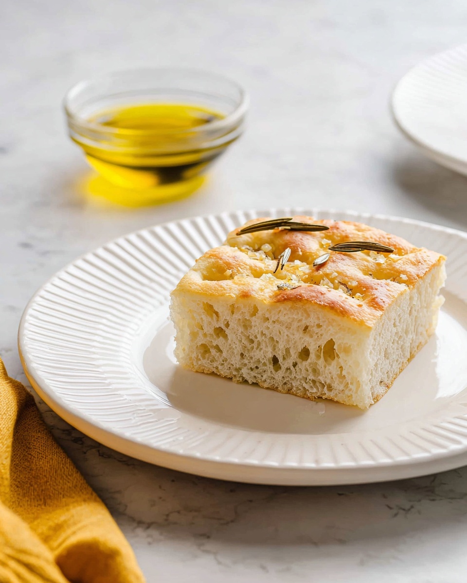 A square piece of soft, light beige focaccia bread with a slightly golden top layer sprinkled with small bits of coarse salt and a few rosemary sprigs is centered on a white plate with raised ridges along the edge. The bread's inside texture looks airy with small holes. Behind it, there is a small clear glass bowl filled with golden olive oil, both placed on a white marbled textured surface. A folded yellow cloth is partially visible on the bottom left corner of the image. Photo taken with an iphone --ar 4:5 --v 7