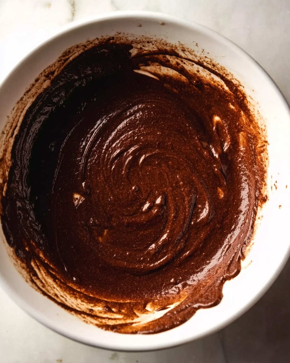 A white bowl filled with thick, dark brown chocolate batter with a smooth, swirled texture on top. Some batter drips over the edge of the bowl onto a white marbled surface below. The rich batter looks creamy with subtle lighter brown streaks mixed in, creating a marbled effect. The edges of the bowl show light splashes of batter, adding to the sense of fresh mixing. The image is close up, focusing on the bowl and batter with soft, natural lighting. Photo taken with an iphone --ar 4:5 --v 7