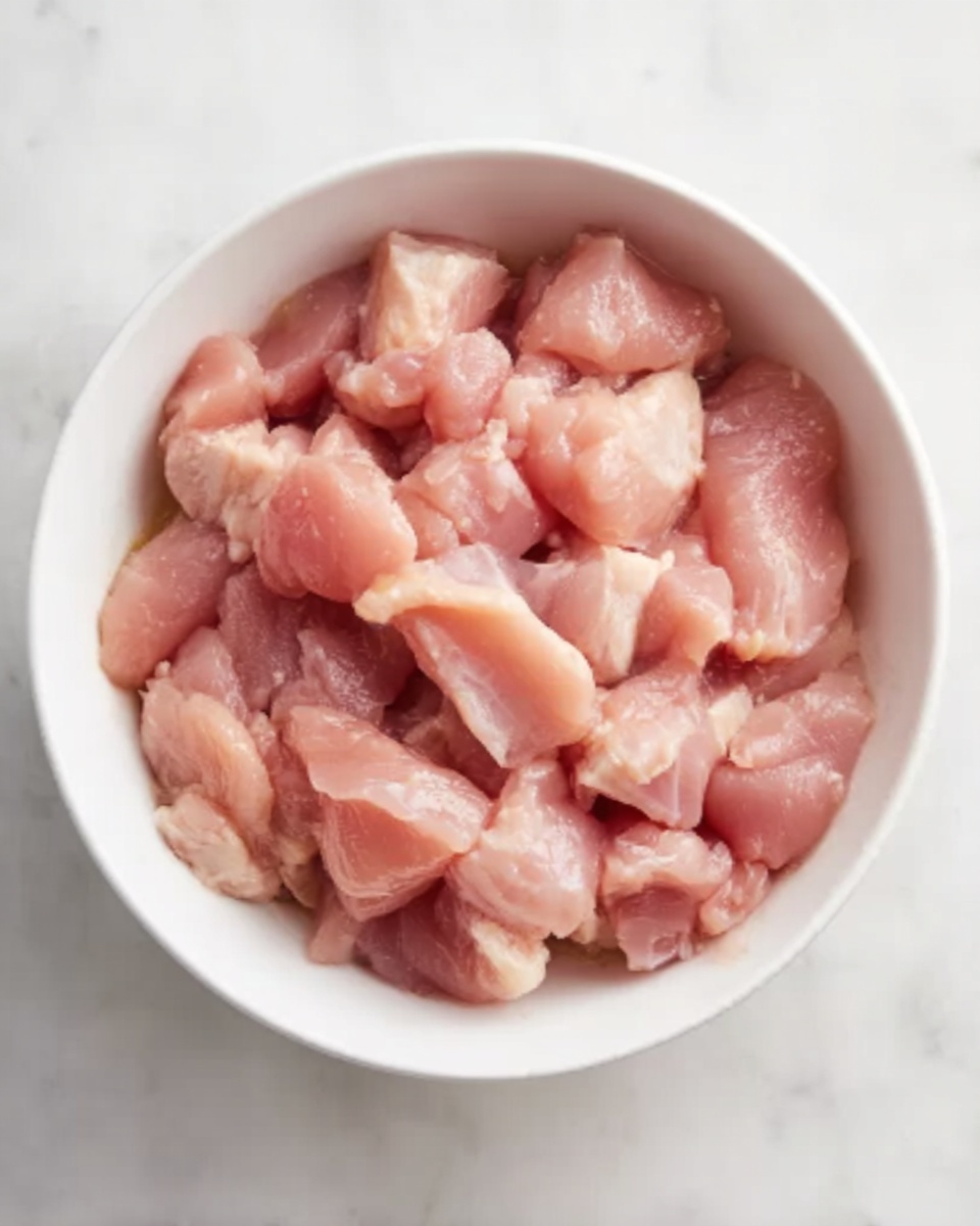 A white bowl filled with small pieces of raw pink chicken, slightly shiny and moist, placed on a white marbled surface. The chicken pieces vary in size and shape, looking fresh and tender. The lighting is soft, showing the texture of the chicken clearly. photo taken with an iphone --ar 4:5 --v 7