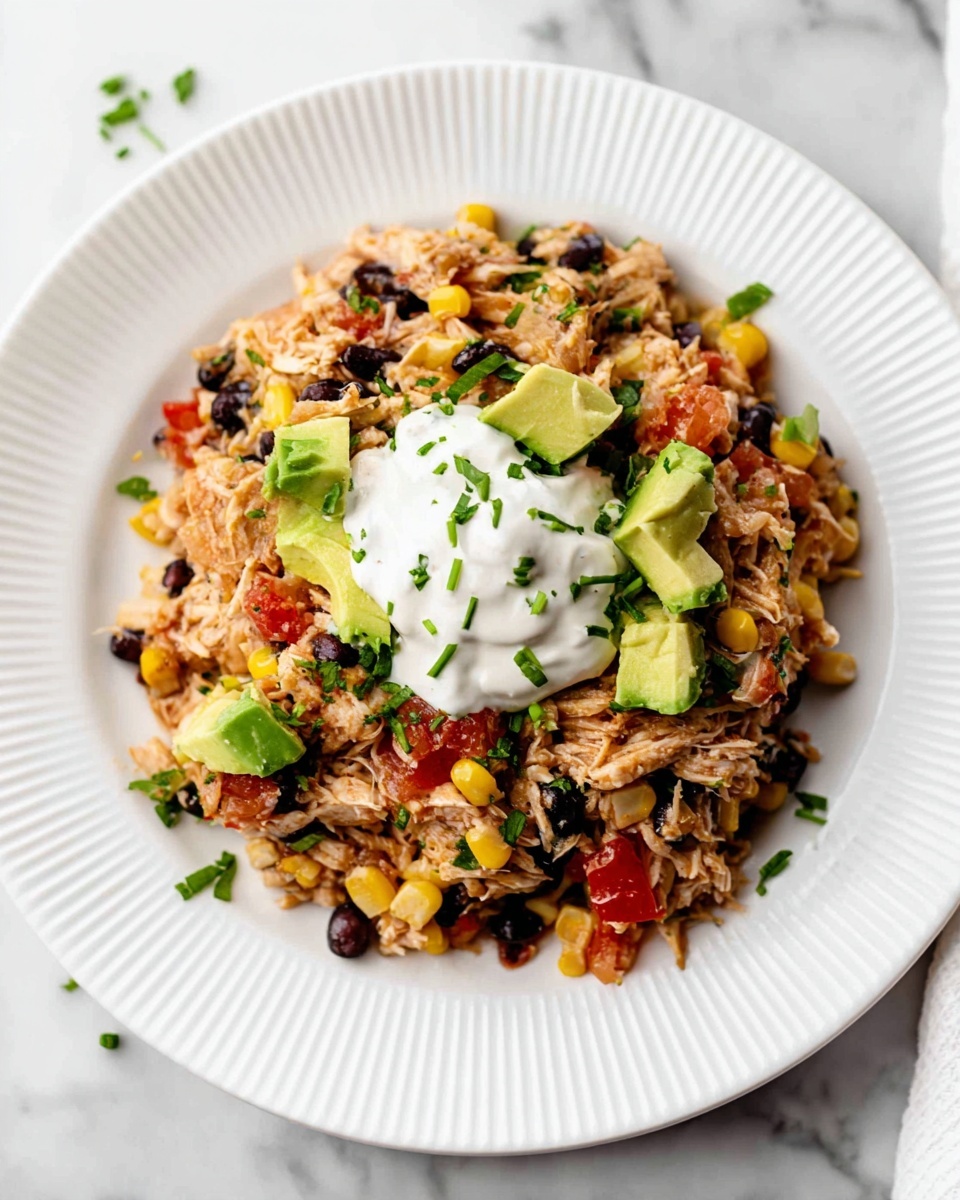 The dish is served on a white plate with a ridged design, placed on a white marbled surface. There is one main layer made of shredded chicken mixed with black beans, yellow corn, and small pieces of red bell pepper, giving a mix of light brown, black, yellow, and red colors with a soft texture. Scattered on top are bright green chunks of avocado and chopped green onions, adding freshness and a slight contrast. In the center of the dish, there is a dollop of thick, creamy white sauce. Small bits of chopped green herbs are sprinkled around the dish for a final touch of color. Photo taken with an iphone --ar 4:5 --v 7