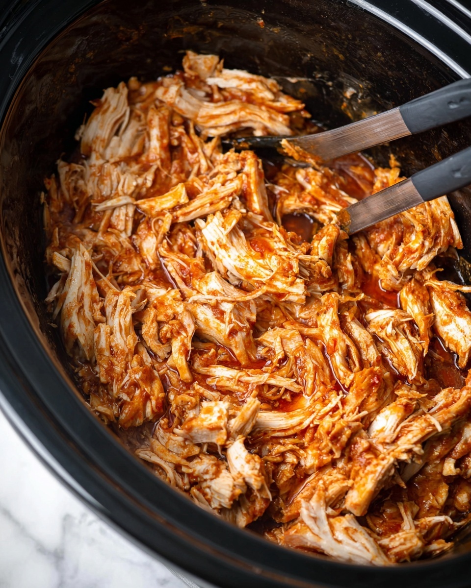 A close-up view of a black slow cooker filled with shredded chicken mixed with a reddish-orange sauce evenly spread over the pieces, showing moist and tender texture with visible strands of meat. The chicken layers are loosely piled within the cooker, and a utensil with dark handles is positioned at the edge, partially in the dish. The background is a white marbled surface. Photo taken with an iphone --ar 4:5 --v 7