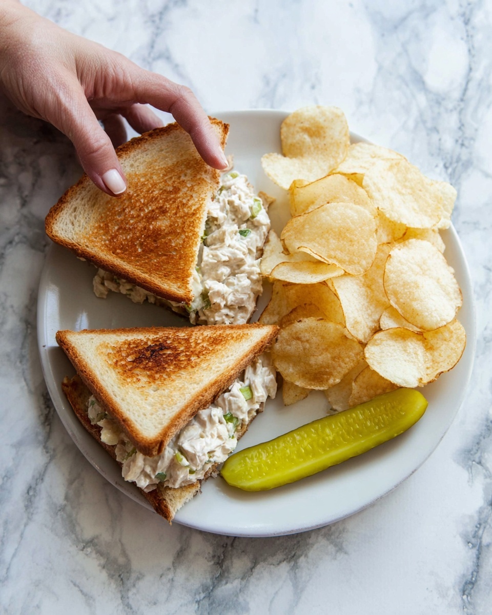 A white round plate holds a sandwich, potato chips, and a pickle slice on a white marbled surface. The sandwich is cut in half diagonally, showing two toasted golden brown triangular bread slices with a creamy light beige chicken salad filling mixed with small green bits inside. The sandwich is positioned on the left side of the plate. A pile of pale golden potato chips with slight brown edges is placed next to the sandwich on the right side. A single bright green pickle slice lies at the bottom edge of the plate, parallel to the sandwich. A woman's hand is touching the sandwich's top left corner. photo taken with an iphone --ar 4:5 --v 7