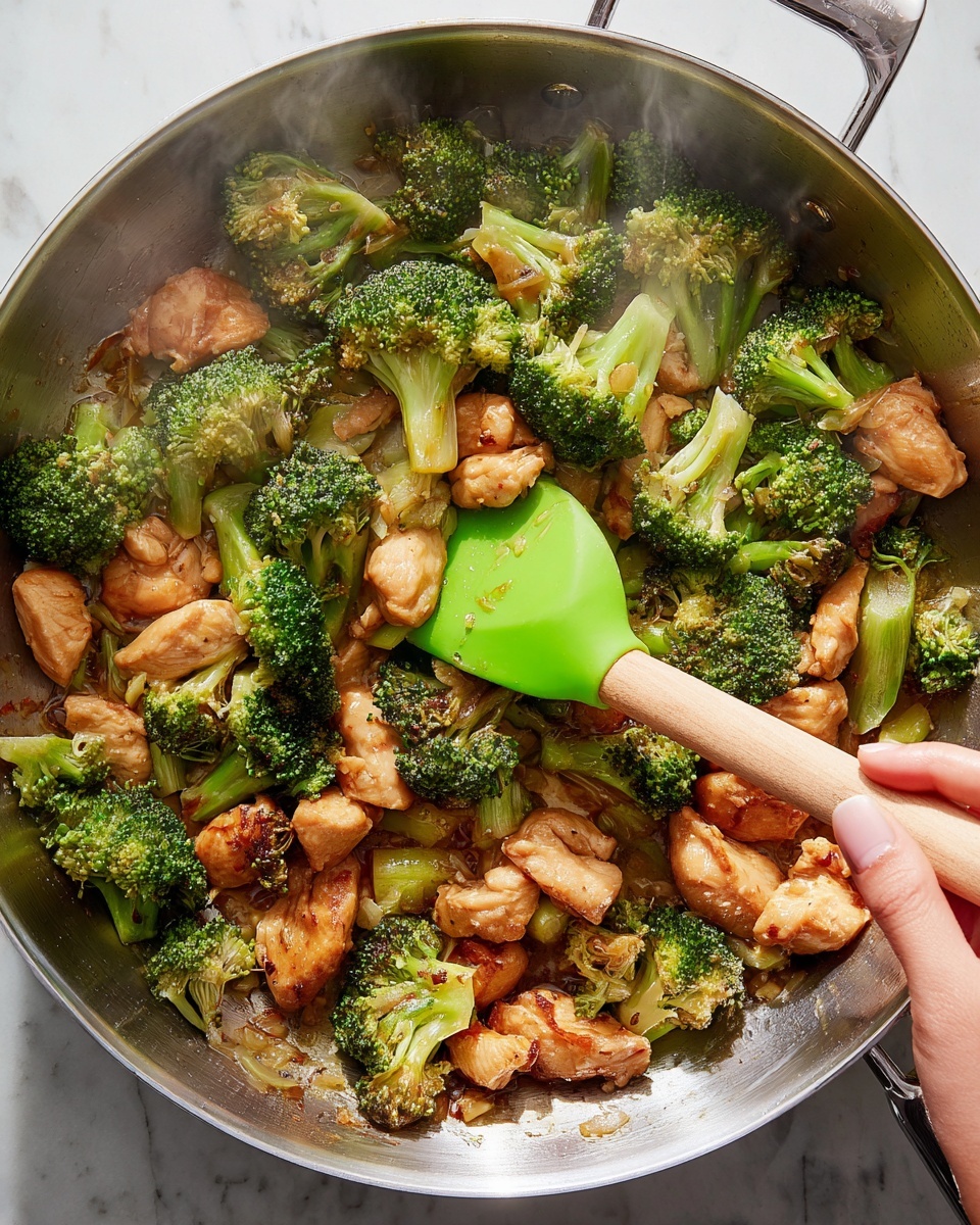 The image shows a large silver pan filled with cooked broccoli and light brown pieces of chicken, all mixed in a slightly shiny sauce. The broccoli is bright green with some parts a little darkened from cooking, while the chicken pieces are golden and slightly crisp on the edges. A green silicone spatula with a wooden handle is stirring the food, held by a woman's hand. The pan is set on a white marbled surface with some steam rising from the food, showing it is hot and fresh. Photo taken with an iphone --ar 4:5 --v 7