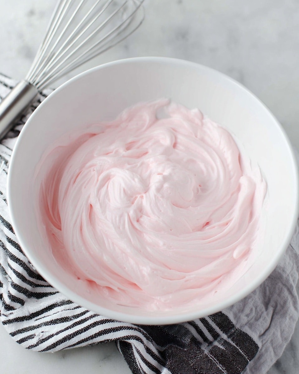 A white bowl filled with light pink whipped cream or frosting, thick and smooth with soft swirls and peaks covering the whole inside of the bowl. The bowl sits on a softly blurred white marbled surface, with a metal whisk and a black-and-white striped cloth partially visible in the lower corner. The texture looks creamy and airy with delicate ridges from stirring, and the pale pink color is even throughout. photo taken with an iphone --ar 4:5 --v 7