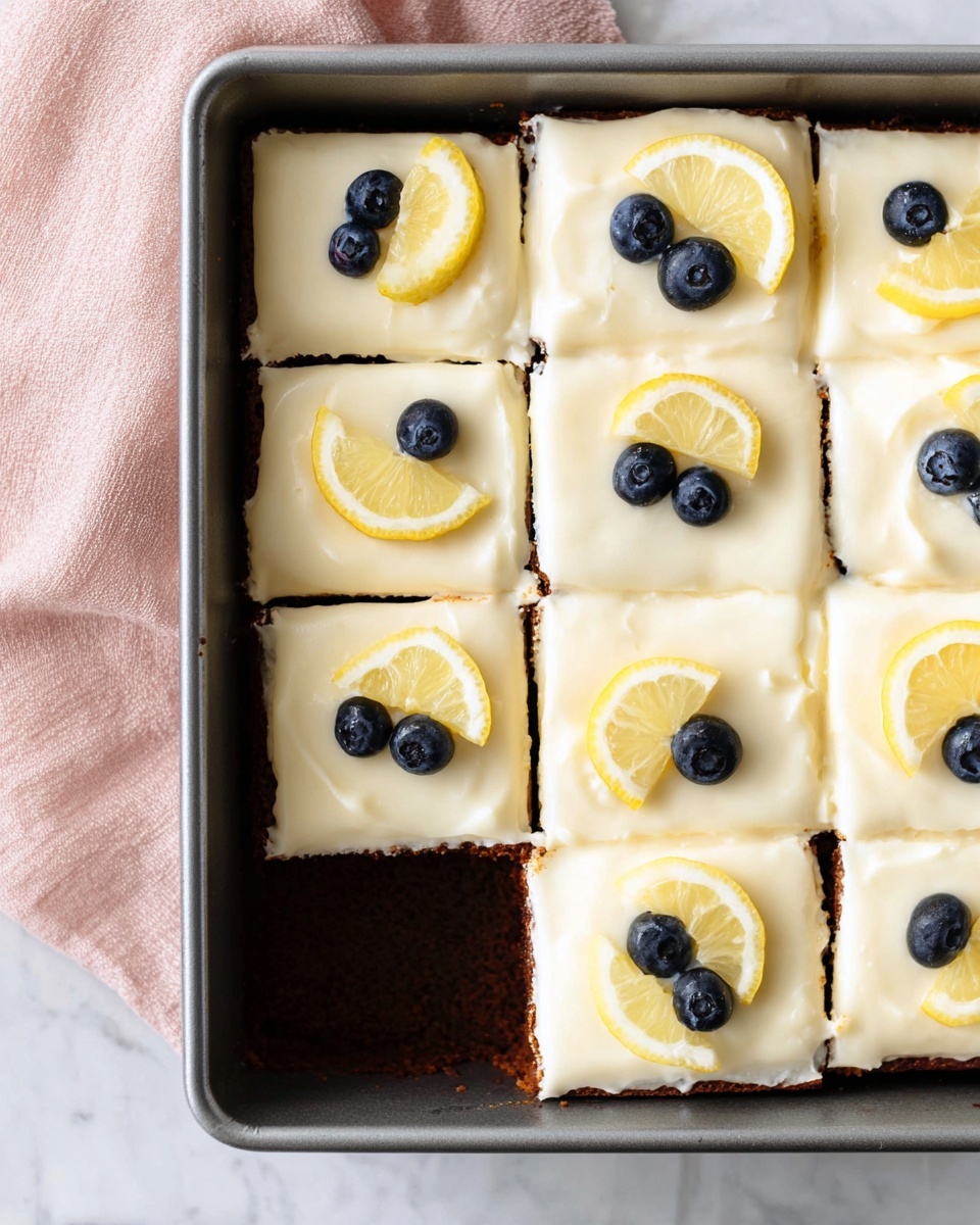 A rectangular cake pan holds nine square pieces of cake with a bottom layer of dark brown cake visible where one piece is missing from the lower left corner. Each piece has a thick, smooth, creamy white frosting layer covering the top. On each frosted square, there is a small, thin lemon wedge positioned near the top edge and two fresh blueberries placed close together just below the lemon slice. The pan sits on a white marbled surface with a soft pink cloth nearby. Photo taken with an iphone --ar 4:5 --v 7