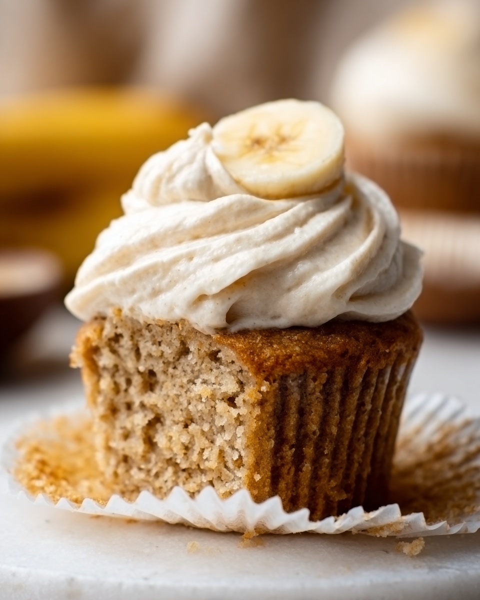 A close-up of a single cupcake with a light brown, soft, and crumbly texture, topped with a thick layer of creamy white frosting that is slightly swirled. A small slice of banana is placed on top of the frosting. The cupcake is in a white paper liner resting on a white marbled surface. In the background, there are blurred objects that add a warm, cozy feel. photo taken with an iphone --ar 4:5 --v 7