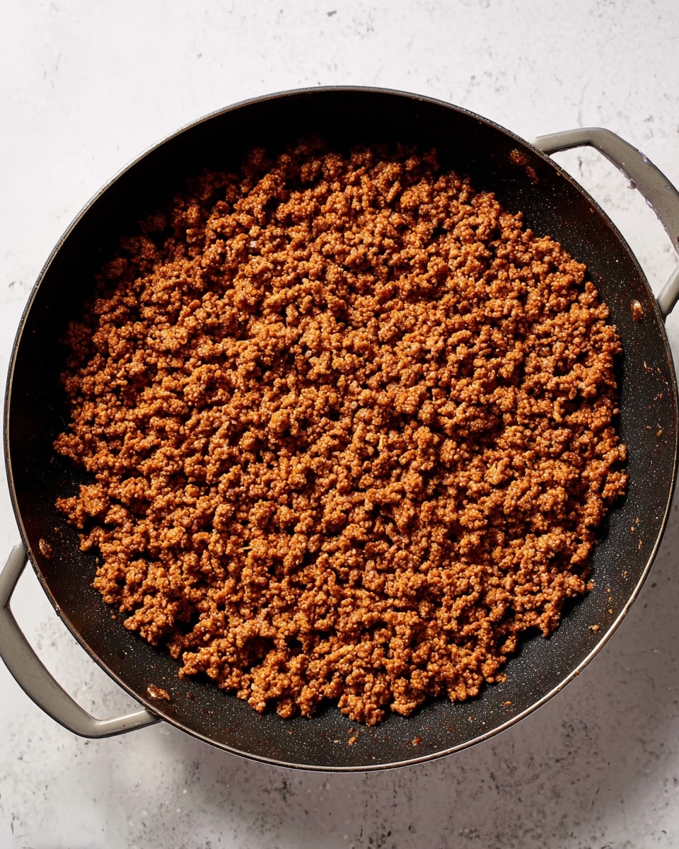 A large black pan with two gray handles holds a layer of cooked browned ground meat spread evenly inside the pan. The meat pieces are small and crumbly with a slightly oily texture, showing a rich brown color with some darker spots. The background surface is a white marbled texture. Photo taken with an iphone --ar 4:5 --v 7