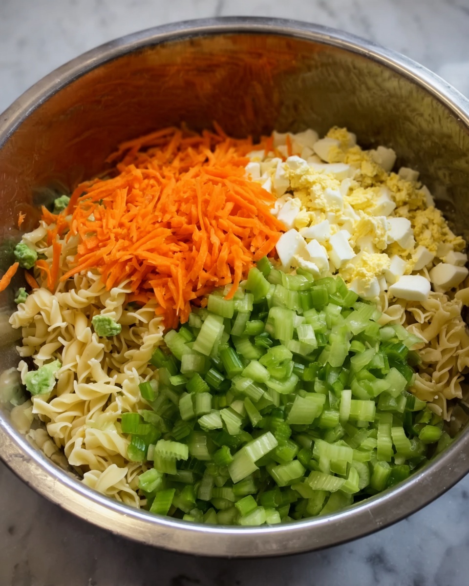 The image shows a large metal bowl filled with a layered salad mix. The first visible layer is small, light beige pasta that fills the base of the bowl. On top of the pasta, there are finely chopped boiled eggs scattered evenly. To the left, there is a bright layer of shredded orange carrots piled neatly. The middle of the bowl has a generous amount of sliced light green celery that covers a big part of the salad. The ingredients all sit together in a shiny, smooth metal bowl, placed on a white marbled surface. Photo taken with an iphone --ar 4:5 --v 7