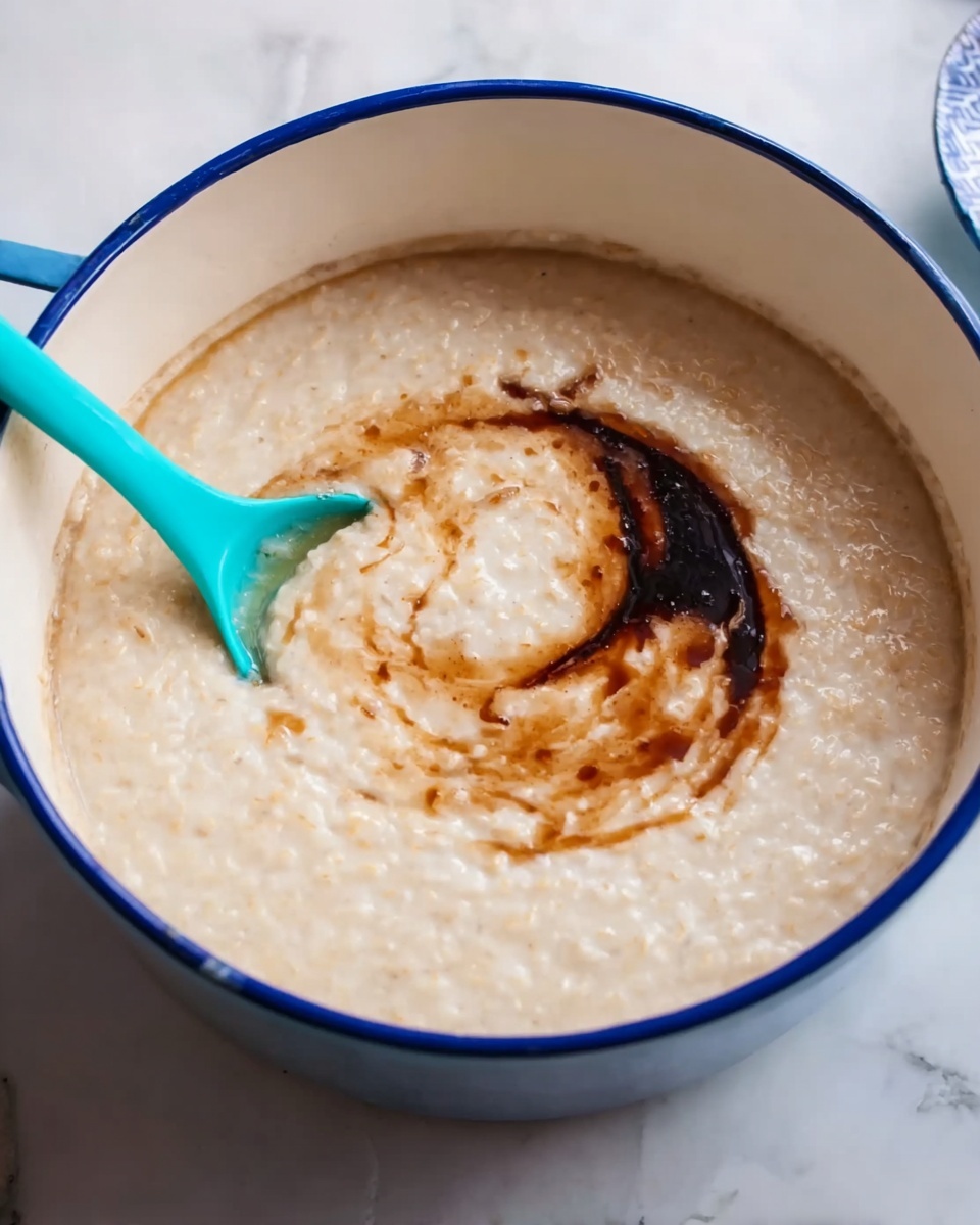 A large white pot with a blue rim is filled with creamy, light beige porridge with a smooth texture. A swirl of dark brown liquid, likely syrup, is added on top near the center. A bright turquoise spoon is partially dipped into the porridge on the left side of the pot. The pot sits on a white marbled surface. photo taken with an iphone --ar 4:5 --v 7
