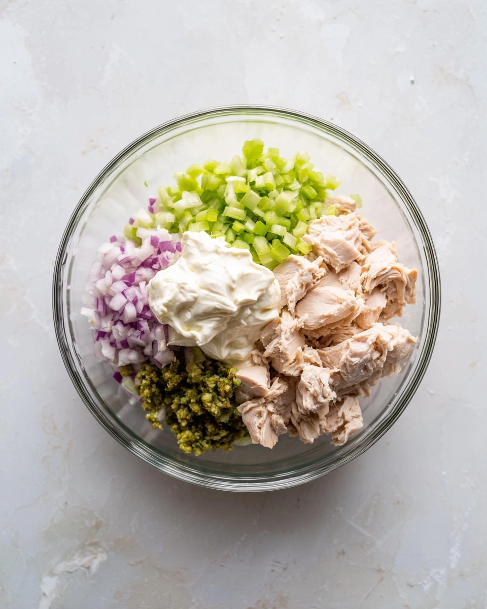 A clear glass bowl holds five distinct piles of ingredients arranged side by side: pale pink chunks of cooked chicken at the center-right, bright white creamy mayonnaise on top of the chicken, finely chopped light green celery at the top left, small pieces of finely chopped purple onion below the celery, and bright green relish at the bottom left near the chicken. The bowl rests on a white marbled surface with subtle gray veins and soft natural light highlights the textures of each ingredient. photo taken with an iphone --ar 4:5 --v 7