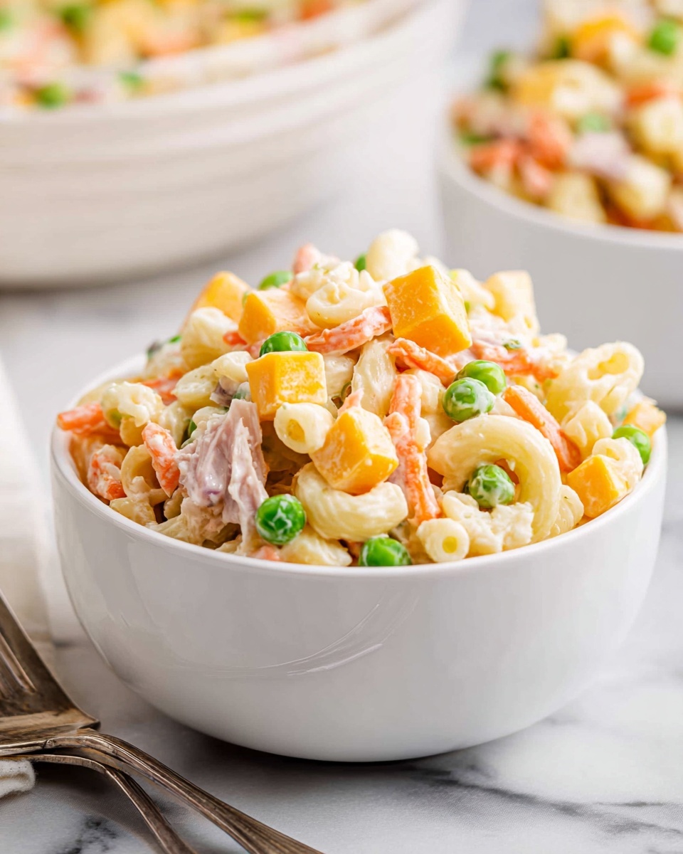 A white bowl filled with a layered salad mix, showing creamy macaroni pasta at the base in pale yellow, with bright orange carrot strips, green peas, and small light yellow egg pieces mixed throughout. Cubes of orange cheese are evenly spread inside, with thin strips of light pink meat or turkey on top. The salad looks creamy with a light dressing, all set on a white marbled surface with a silver fork on the side. In the blurry background, another white bowl of the same salad is visible. Photo taken with an iphone --ar 4:5 --v 7