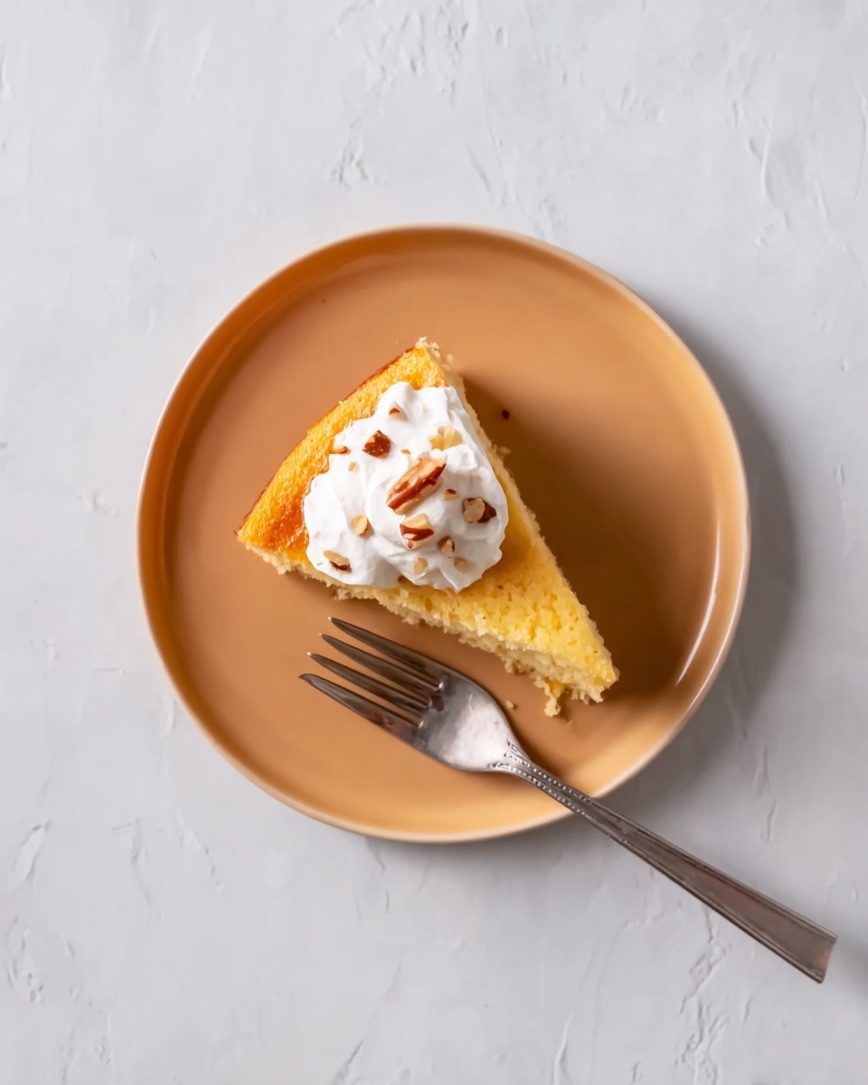 A single slice of yellow cake sits in the middle of a white plate with a smooth, slightly shiny surface. The cake slice is topped with a dollop of white whipped cream that looks soft and fluffy. Small brown bits, like nuts, are sprinkled on top of the whipped cream. To the right of the cake slice lies a silver fork, placed at a slight angle with its handle extending beyond the plate's edge. The plate rests on a white marbled surface showing faint gray veins and subtle texture. photo taken with an iphone --ar 4:5 --v 7