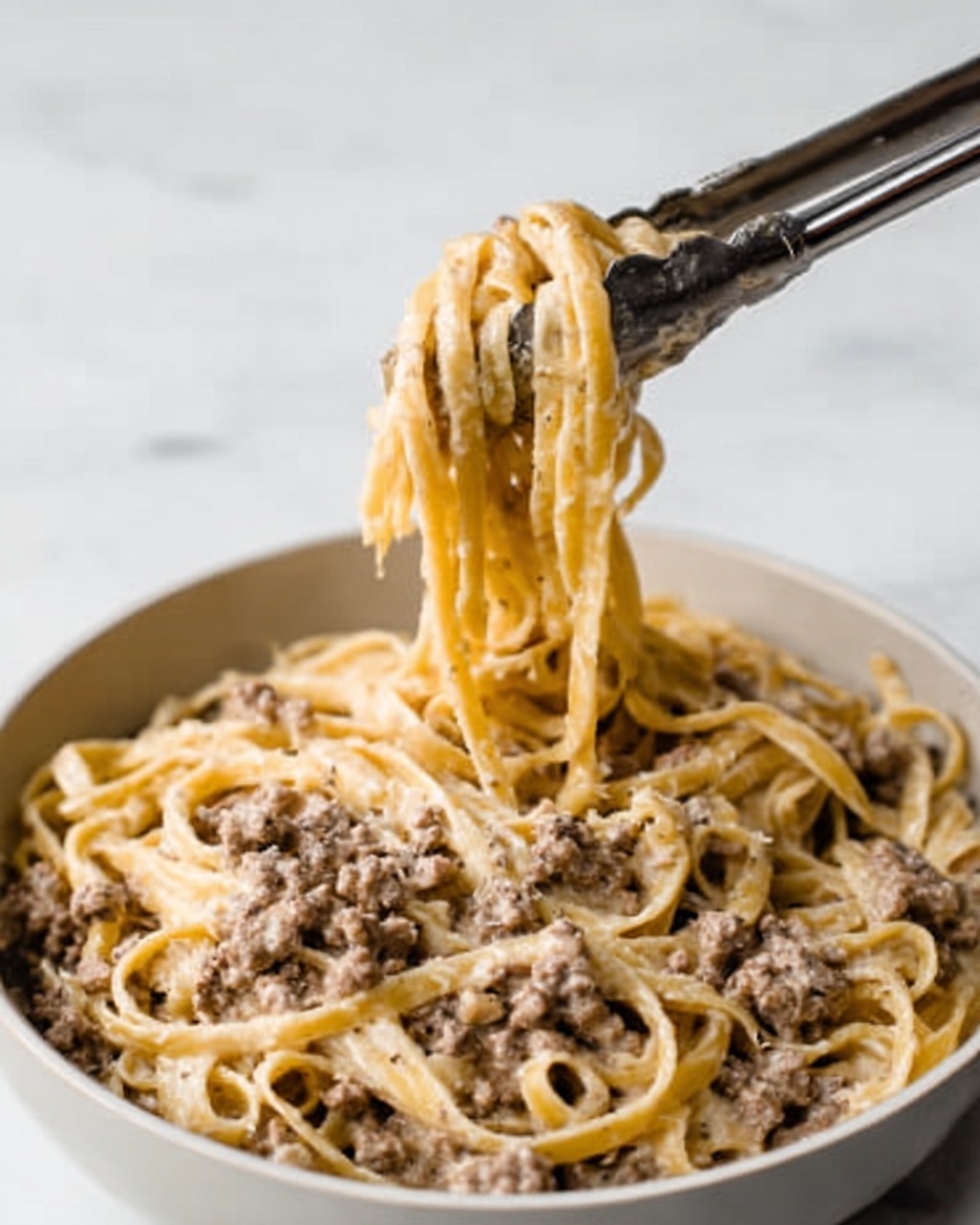 The image shows a close-up of a bowl of creamy pasta with ground meat. The pasta is long and flat, covered in a light beige sauce mixed well with brownish ground meat pieces. A pair of silver tongs lift a tangled bunch of the pasta from the white bowl, showing the sauce clinging to the noodles. The bowl sits on a white marbled surface, and the background is soft and out of focus. photo taken with an iphone --ar 4:5 --v 7