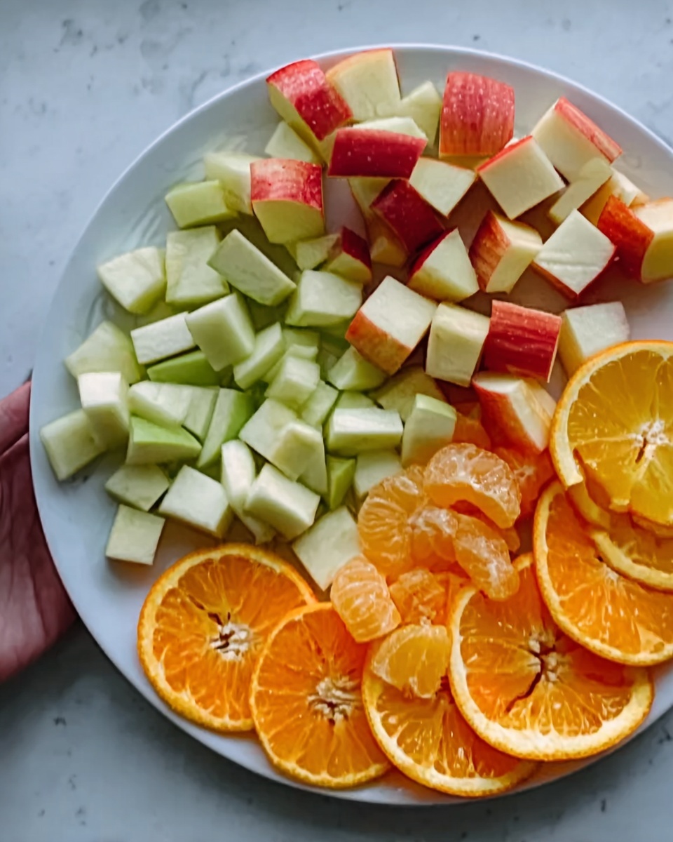 The image shows a white plate with three kinds of sliced fruits neatly arranged. At the top part of the plate are red apple pieces, each cut into small chunks with green skin showing. Below the apples, green apple slices are arranged in small cubes with their skin intact. Towards the bottom of the plate, round, thin slices of orange and yellow citrus fruits are layered, showing bright orange and yellow colors with the soft, juicy texture visible. The plate is placed on a white marbled surface, and a woman's hand is slightly touching the plate edge. photo taken with an iphone --ar 4:5 --v 7