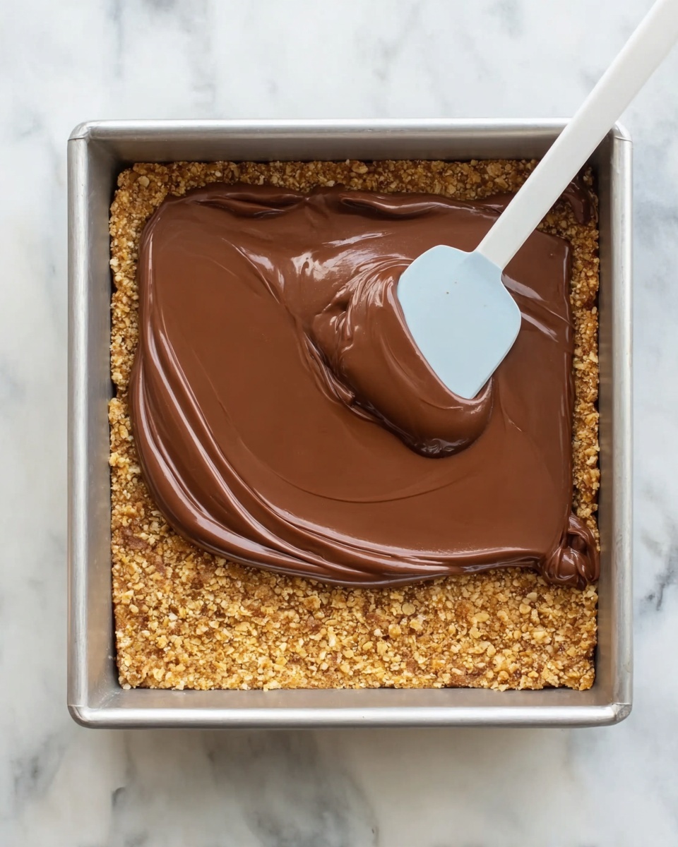 The image shows a square metal baking pan with two layers of a dessert. The bottom layer is made of a golden brown crumbly base, which looks like crushed biscuits or oats packed evenly. The top layer is a thick, smooth, shiny milk chocolate spread being evenly smoothed over the base with a white spatula. The background is a white marbled surface. Photo taken with an iphone --ar 4:5 --v 7
