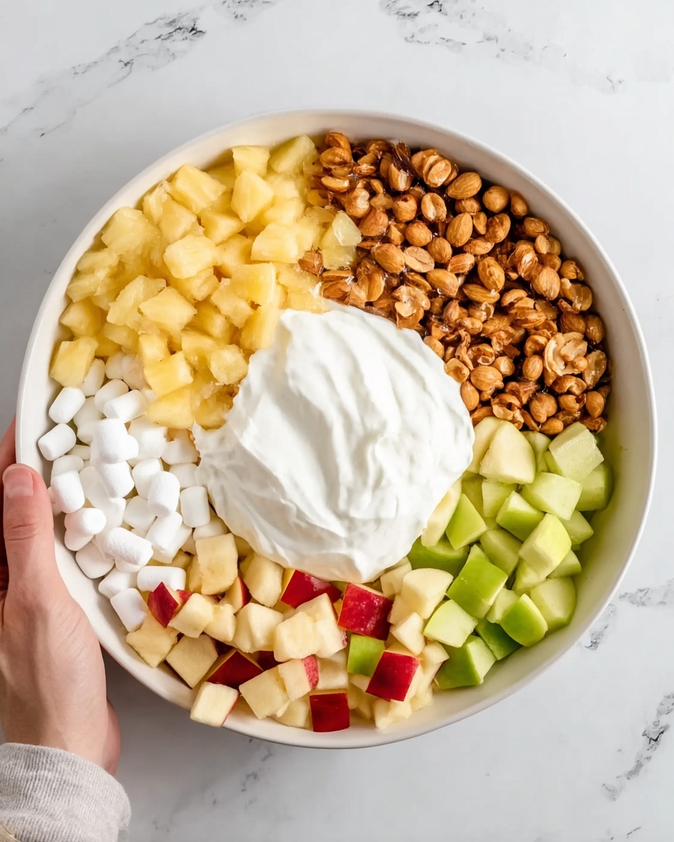 A white bowl filled with five different layers of food arranged separately in sections, placed on a white marbled surface. Starting from the top left, there is a layer of small, yellow pineapple chunks. Next to it on the top right are golden-brown roasted peanuts. Below the peanuts on the right side, there is a layer of small, white mini marshmallows. In the bottom right section, there are small pieces of red-skinned apple with light yellow flesh. Opposite them on the bottom left side are small green apple chunks with skin on. In the center of the bowl, a large dollop of thick white creamy yogurt sits over the middle of the sections, partially covering some of the fruits and nuts. A woman's hand is reaching toward the bowl from the left side. Photo taken with an iphone --ar 4:5 --v 7