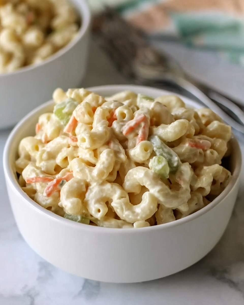 The image shows a small white round bowl filled with creamy macaroni salad. The salad has three main layers: a base of pale yellow cooked elbow macaroni, mixed evenly with small bits of light orange carrot pieces and pale green celery pieces throughout. The macaroni and the vegetables are coated in a thick, smooth, creamy dressing that gives a slightly glossy texture. The bowl is placed on a soft white marbled background, and in the background, there is a larger white bowl with more macaroni salad slightly blurred. A silver fork is also partially visible near the bowl. photo taken with an iphone --ar 4:5 --v 7