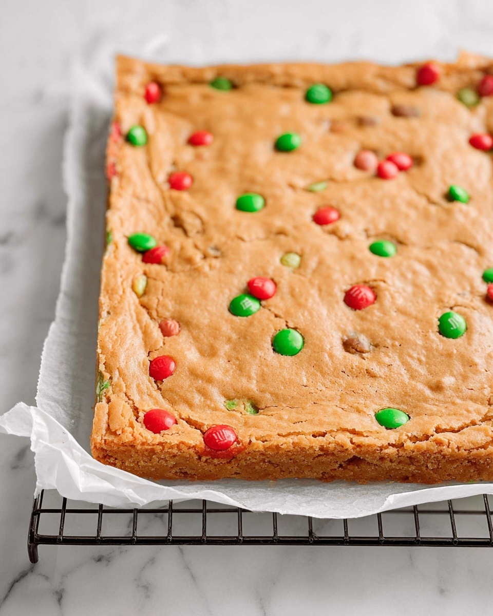 A rectangular baked blondie bar with a light golden brown top layer that looks soft and slightly cracked, embedded with scattered red and green candy-coated chocolates throughout the surface, showing hints of candies inside the edges. The blondie rests on a white parchment paper, which sits on a black metal cooling rack, all set against a white marbled background. photo taken with an iphone --ar 4:5 --v 7