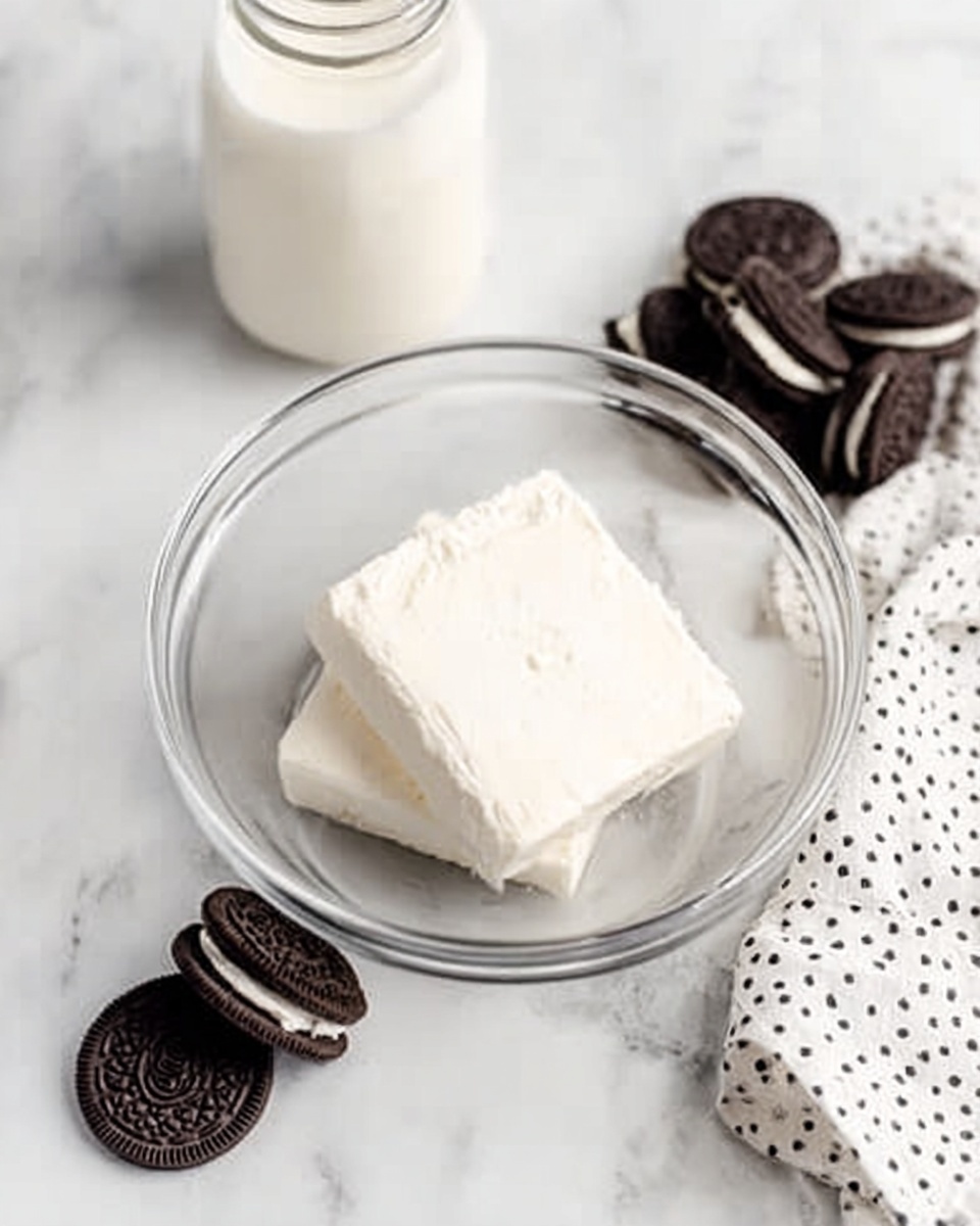 A clear glass bowl on a white marbled surface holds two thick, white blocks of cream cheese stacked one on top of the other in the center. To the left of the bowl is a glass jar filled with milk, and beyond it, a small pile of whole chocolate sandwich cookies is placed, dark brown with creamy white filling visible at the edges. On the right side of the bowl, three separated chocolate sandwich cookies lie flat, showing the dark cookie outer and white filling inside. A white cloth with black polka dots is partially visible on the lower right corner of the image. Photo taken with an iphone --ar 4:5 --v 7