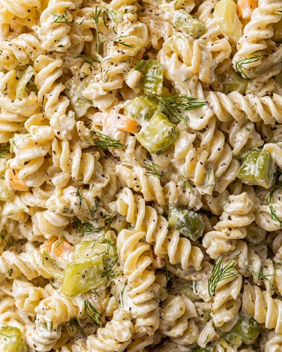 A clear glass bowl filled with creamy pasta salad made of spiral-shaped rotini noodles. The salad has several layers of textured creamy dressing that coats the noodles evenly. Mixed throughout are small green pickle chunks scattered on the top surface, adding color contrast. There are tiny bits of fresh green herbs sprinkled on top, giving a fresh look. The bowl rests on a white marbled surface, with a green sprig of dill and a piece of wood placed nearby, adding natural decoration. photo taken with an iphone --ar 4:5 --v 7