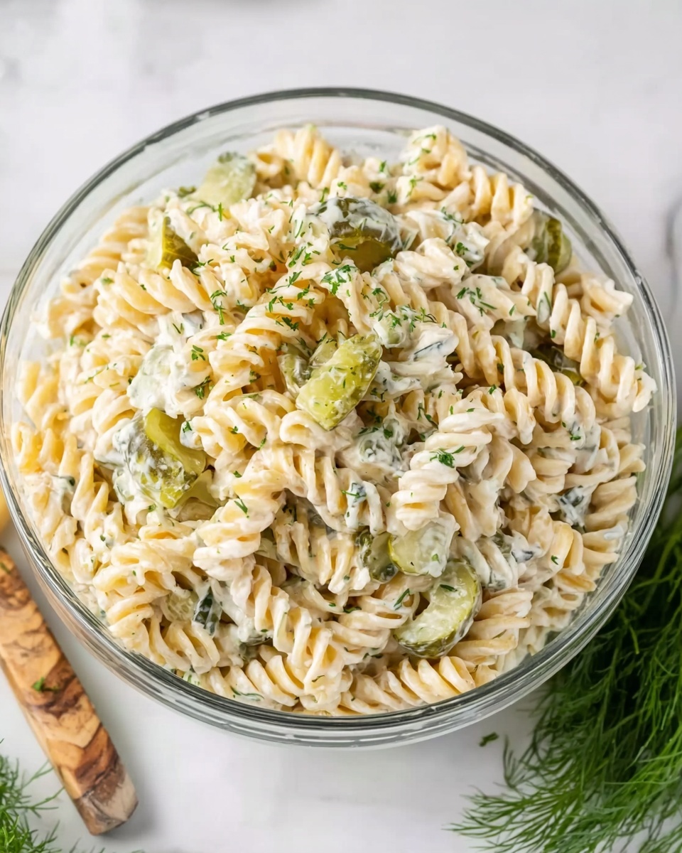 This image shows a close-up view of creamy rotini pasta salad with a single layer of spiral-shaped pasta in light beige, mixed with diced light green pickles and small chunks of pale orange cheese. The salad is sprinkled with fresh green dill and small bits of black pepper, all on a white marbled surface. The creamy dressing coats the pasta and other ingredients evenly, giving a smooth, slightly shiny texture. Photo taken with an iphone --ar 4:5 --v 7