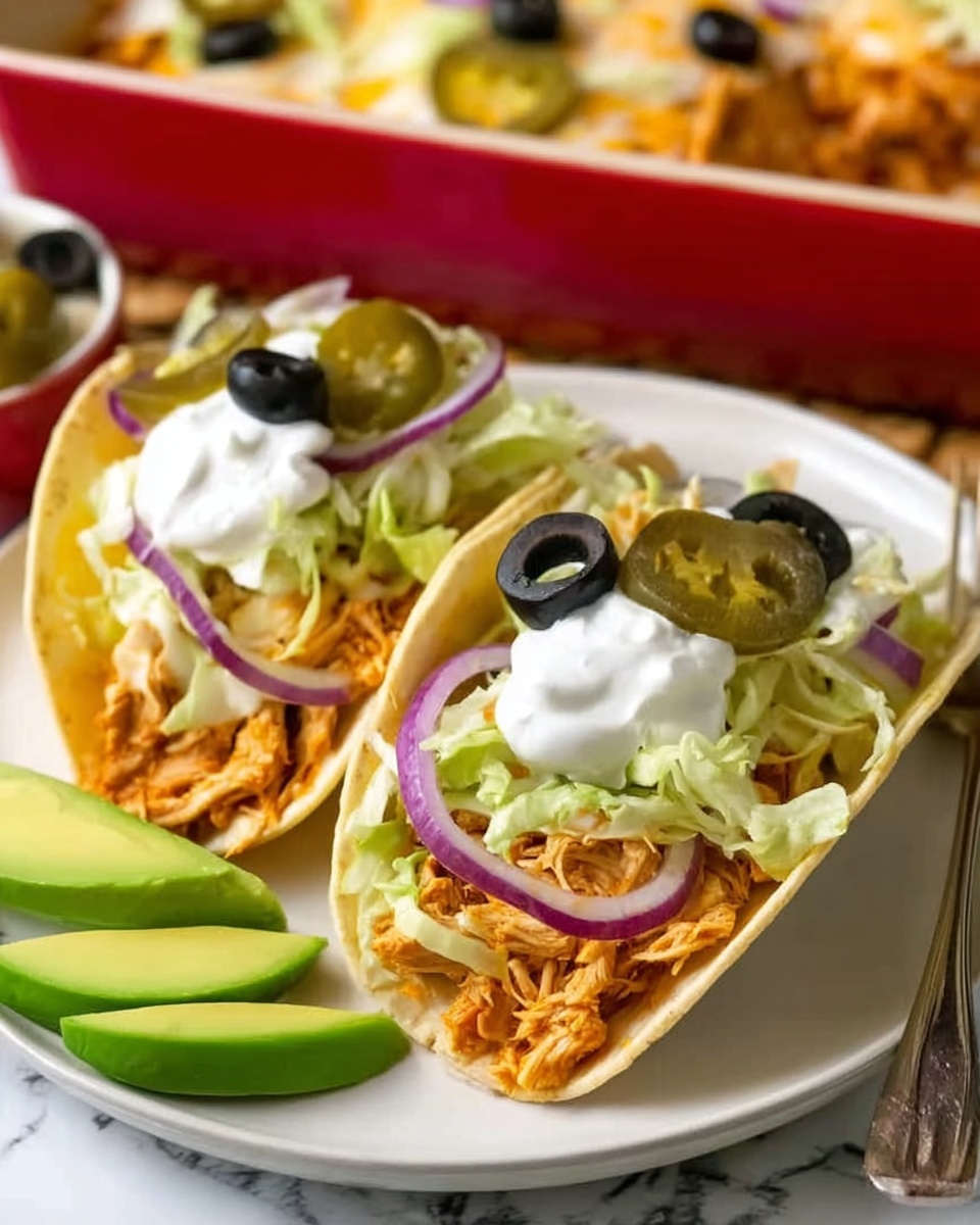 The image shows two tacos placed on a white plate with a white marbled surface underneath. Each taco has three visible layers: a soft beige tortilla filled with shredded orange chicken, topped with light green shredded lettuce, thin slices of purple onion, black olive rings, and a few bright green pickled jalapeño slices. Each taco is finished with two dollops of white sour cream on top. On the plate near the tacos are three slices of fresh avocado with light green flesh. In the background, there is a red casserole dish partially visible, containing more shredded chicken. The photo was taken with an iphone --ar 4:5 --v 7
