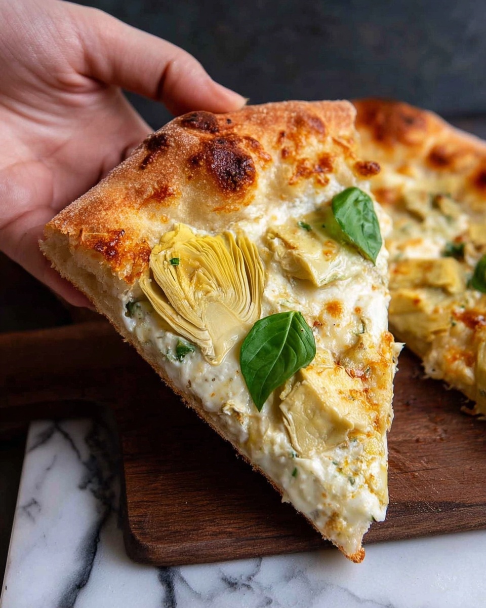 A slice of pizza is shown with a golden brown crust that looks crisp and thick. The pizza has one main layer of melted white cheese spread evenly on the dough. On top of the cheese layer, there are light yellow artichoke pieces and a few small green basil leaves scattered. The edges of the cheese layer have browned spots, adding a toasted texture. A woman's hand is holding the slice gently from the crust. The pizza is placed on a dark wooden surface with a white marbled texture nearby. The photo taken with an iphone --ar 4:5 --v 7