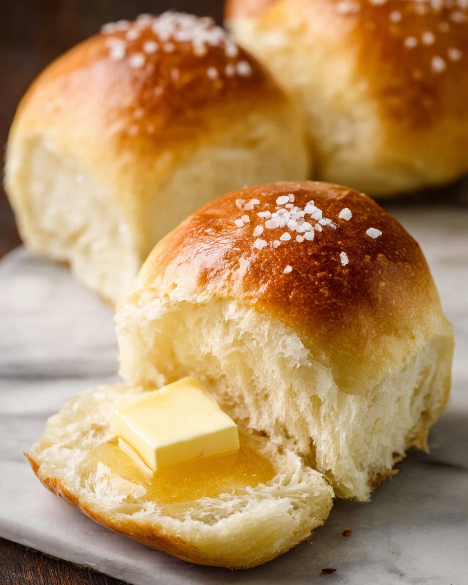 The image shows a soft bread roll with a golden brown top sprinkled with coarse salt. The roll is split open, revealing a light, fluffy white inside. On the inside bottom layer of the roll, there is a square pat of pale yellow butter with a small drizzle of honey on top. In the background, two more whole rolls with shiny golden crusts can be seen on a white marbled surface. photo taken with an iphone --ar 4:5 --v 7