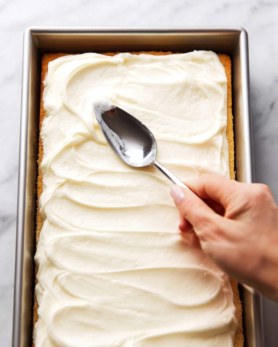 A close-up image shows a rectangular pastry with one golden brown layer covered by a thick, creamy white layer spread evenly on top. A woman's hand is holding a silver spoon, gently smoothing the creamy layer on the pastry corner. The pastry and cream are inside a metal baking pan, and the background is a white marbled texture. Photo taken with an iphone --ar 4:5 --v 7