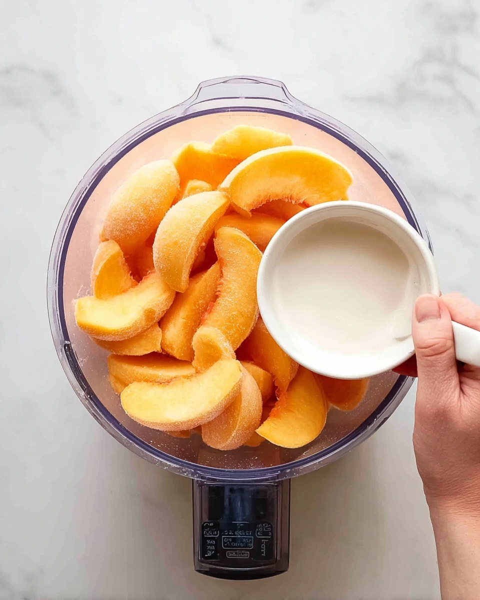 A clear food processor bowl filled with bright orange peach slices sitting on a white marbled surface. A woman's hand holds a small white bowl filled with a smooth white liquid, ready to pour it into the food processor. The peach slices are roughly in a single layer inside the bowl, showing their soft texture and slightly frosted appearance. The overall look is clean and fresh, with soft natural lighting highlighting the vibrant color of the peaches. Photo taken with an iphone --ar 4:5 --v 7