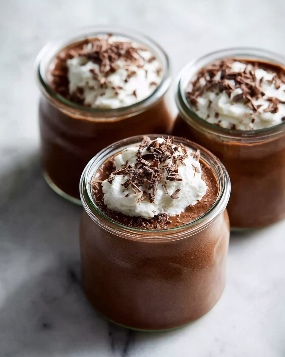 A close-up top view of a glass jar filled with rich chocolate mousse that has a smooth, slightly textured surface. On one side, a thick layer of white whipped cream sits, topped with thin, irregular dark chocolate shavings. Some chocolate shavings are scattered on the whipped cream and mousse, adding contrast to the smooth textures. The jar is placed on a white marbled surface, enhancing the rich colors of the dessert. photo taken with an iphone --ar 4:5 --v 7